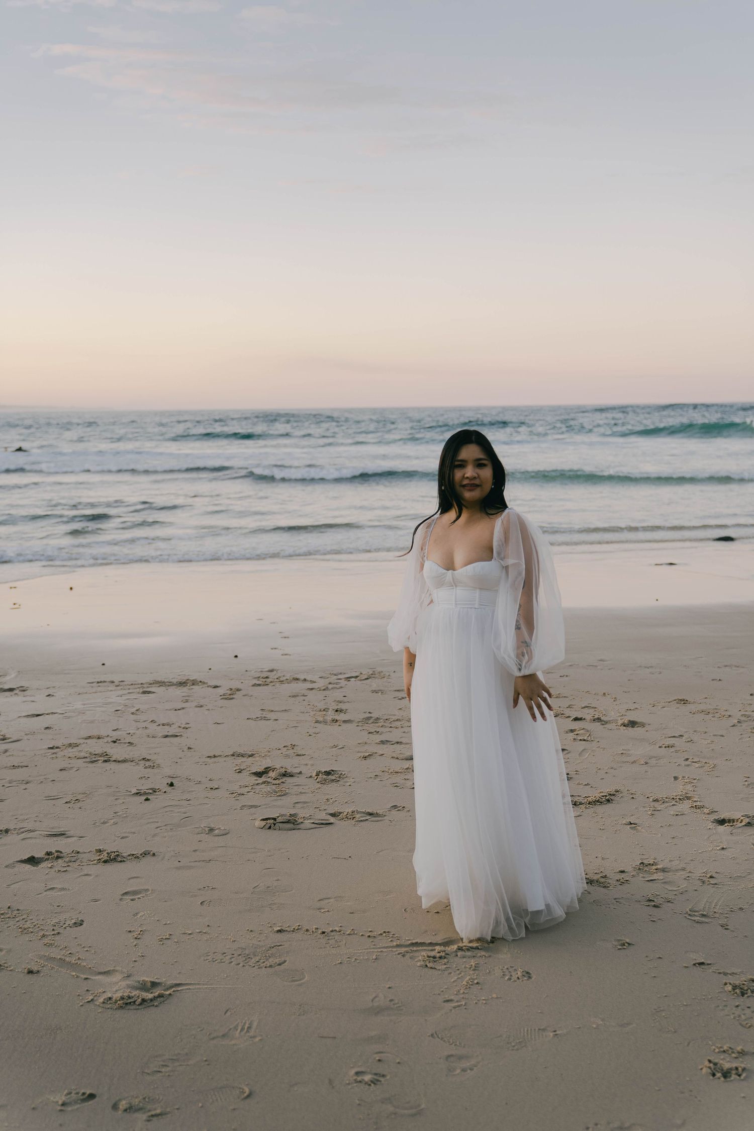 A person in a flowing white dress stands on a sandy beach at sunset with ocean waves in the background.