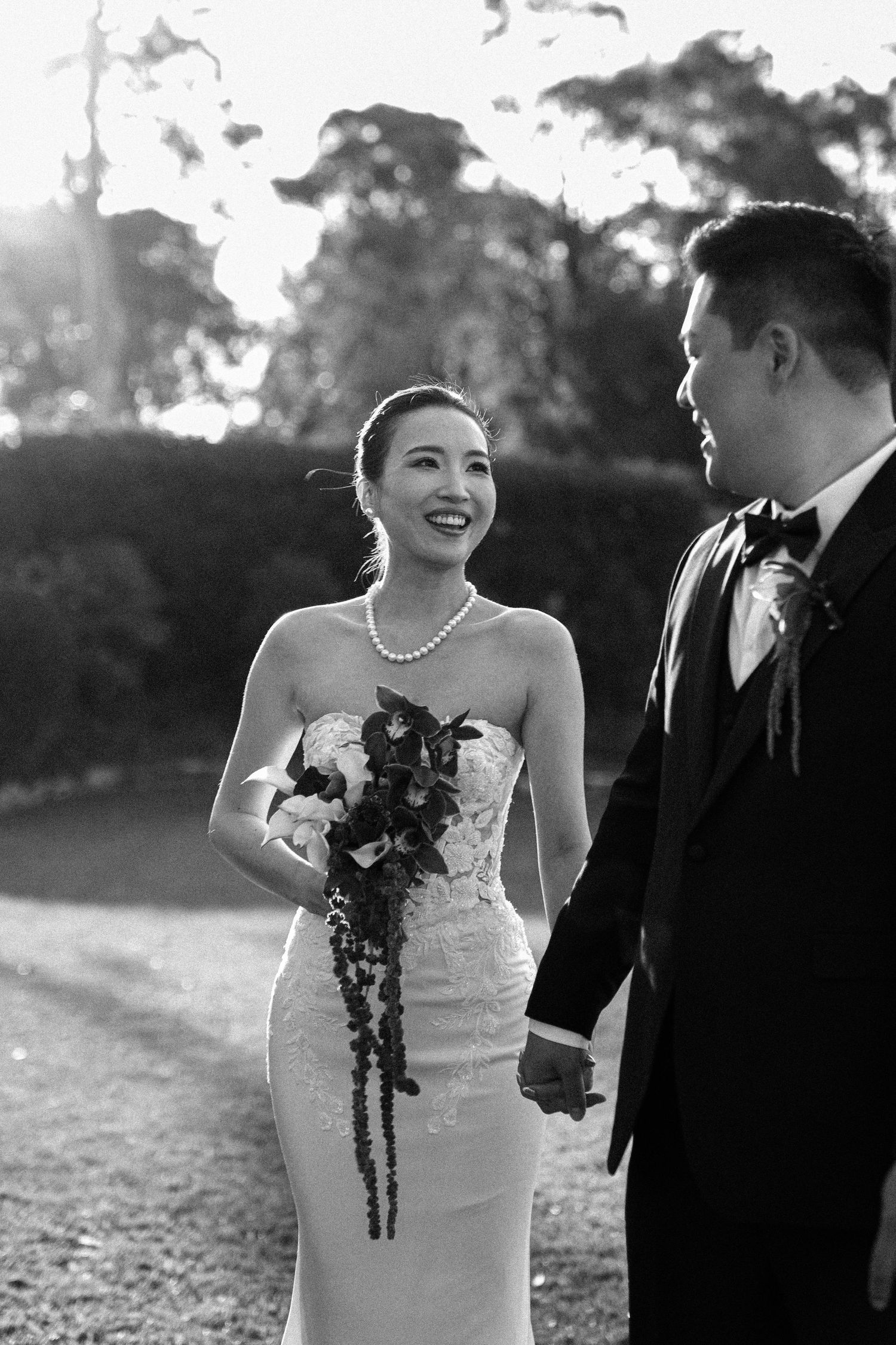 Black and white wedding photo of a couple sharing a joyful moment outdoors with sunlight filtering through trees.