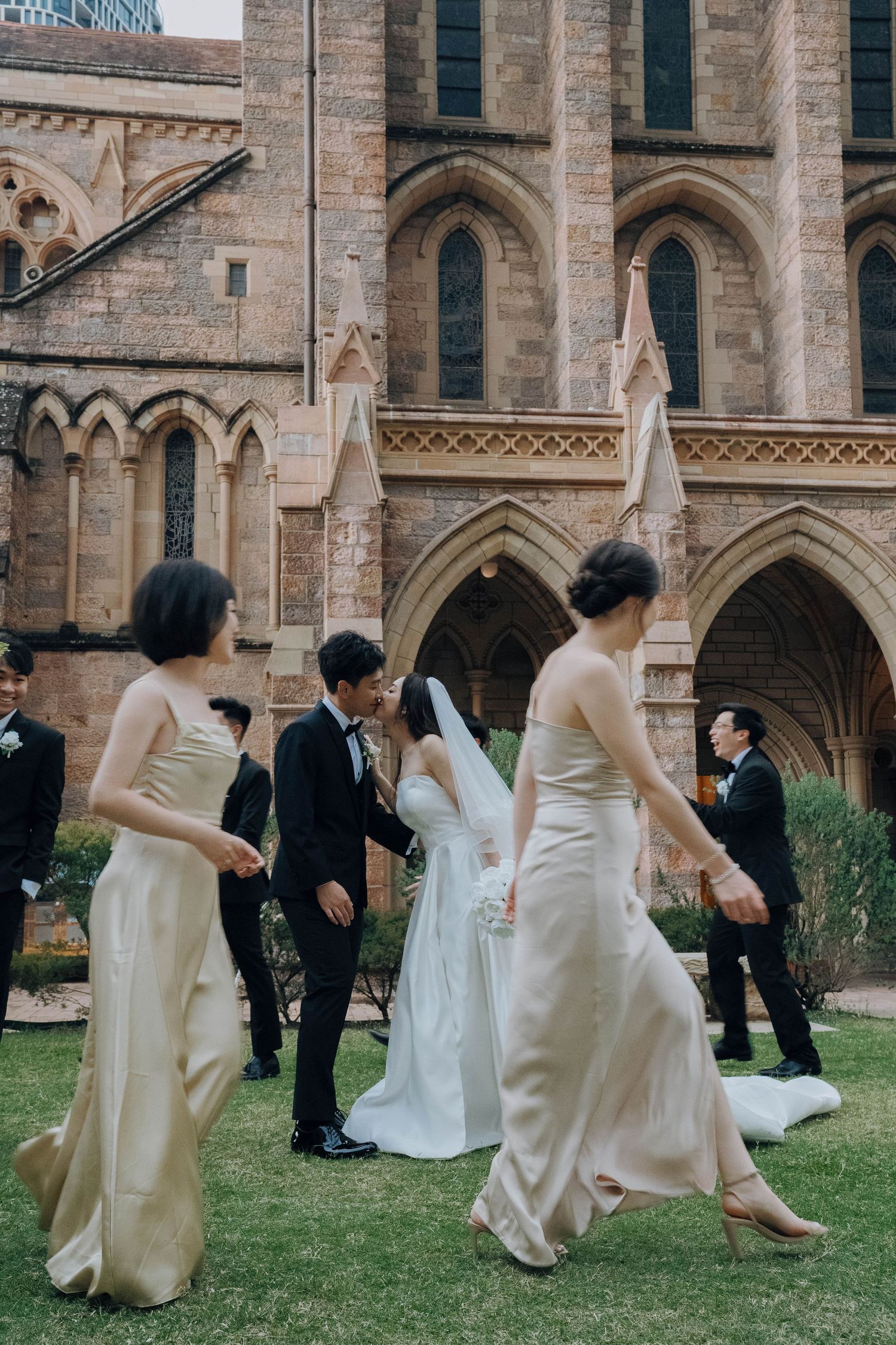 Wedding party in formal attire poses playfully on grass outside a Gothic stone church building.