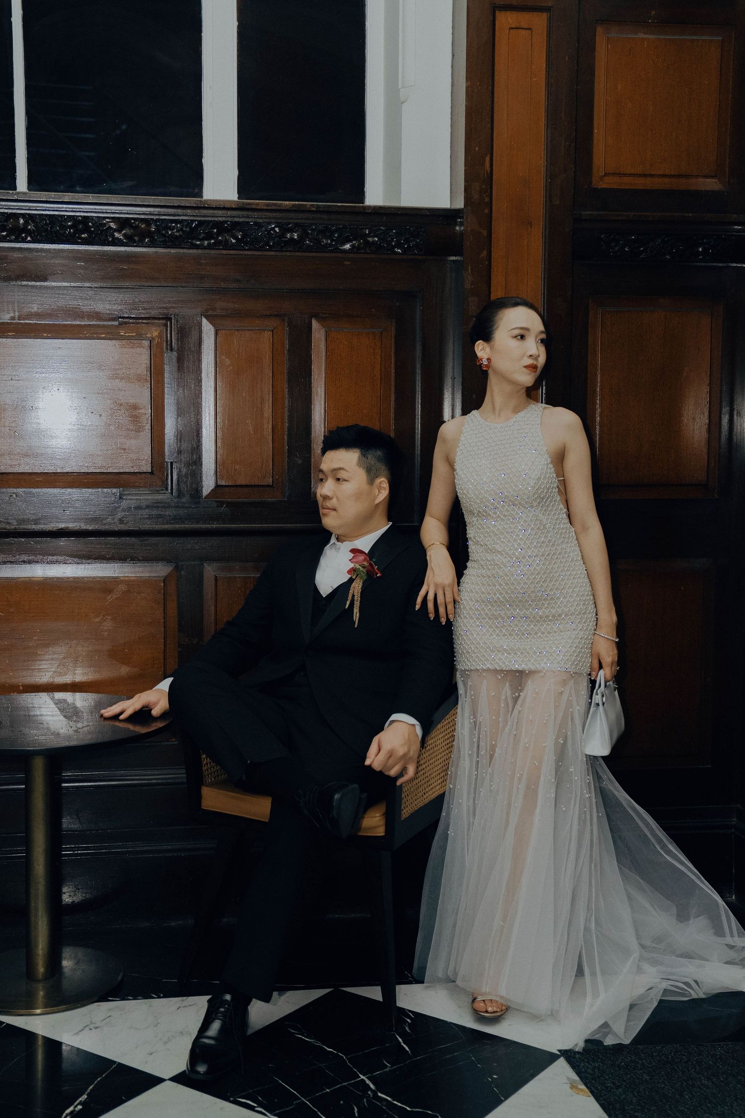 A couple in formal attire poses together in an elegant interior setting with dark wood paneling.