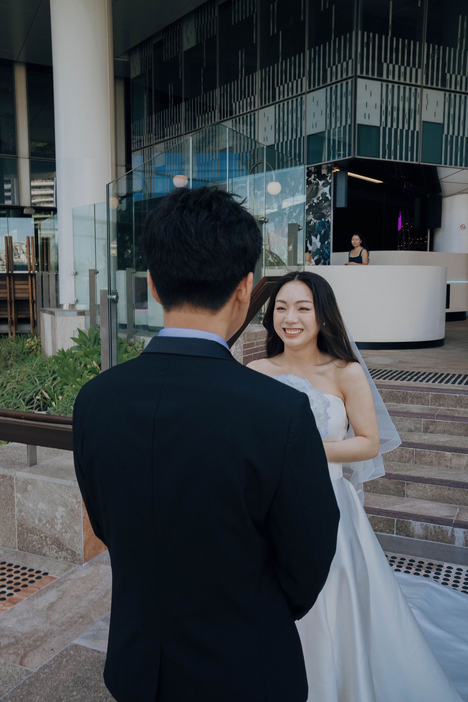 A couple in wedding attire share a joyful moment on modern concrete steps with glass railings in an urban setting.