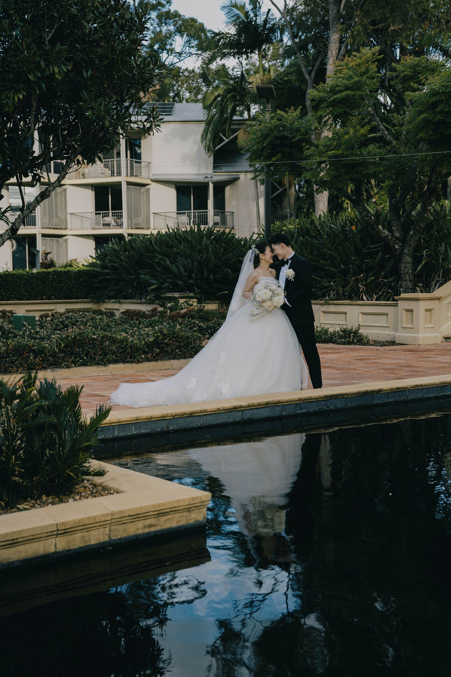 A romantic wedding couple poses along a waterfront boardwalk surrounded by lush tropical landscaping.