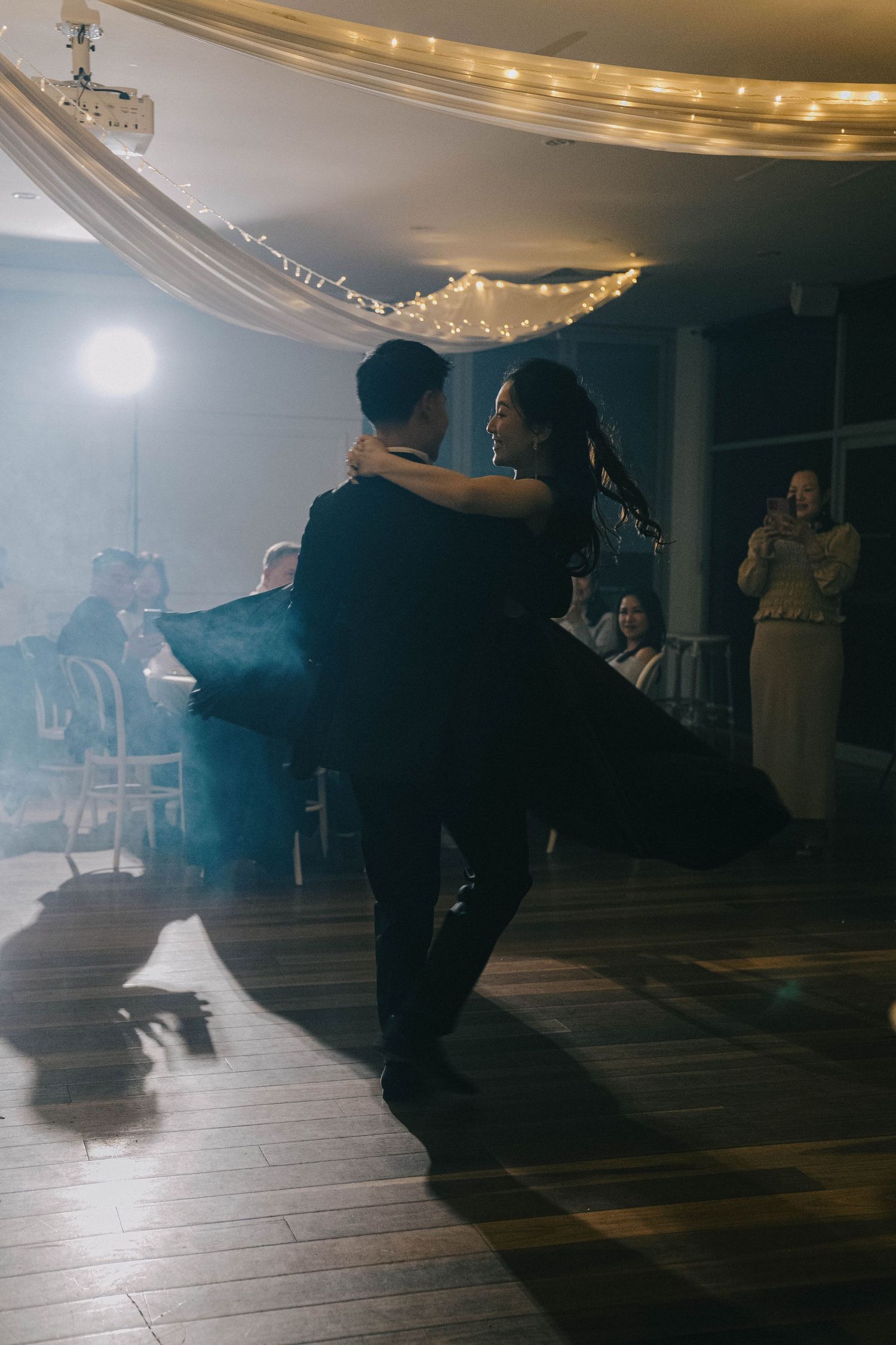 A series of dark silhouettes perform dance movements on a dimly lit wooden floor beneath elegant ceiling lights.