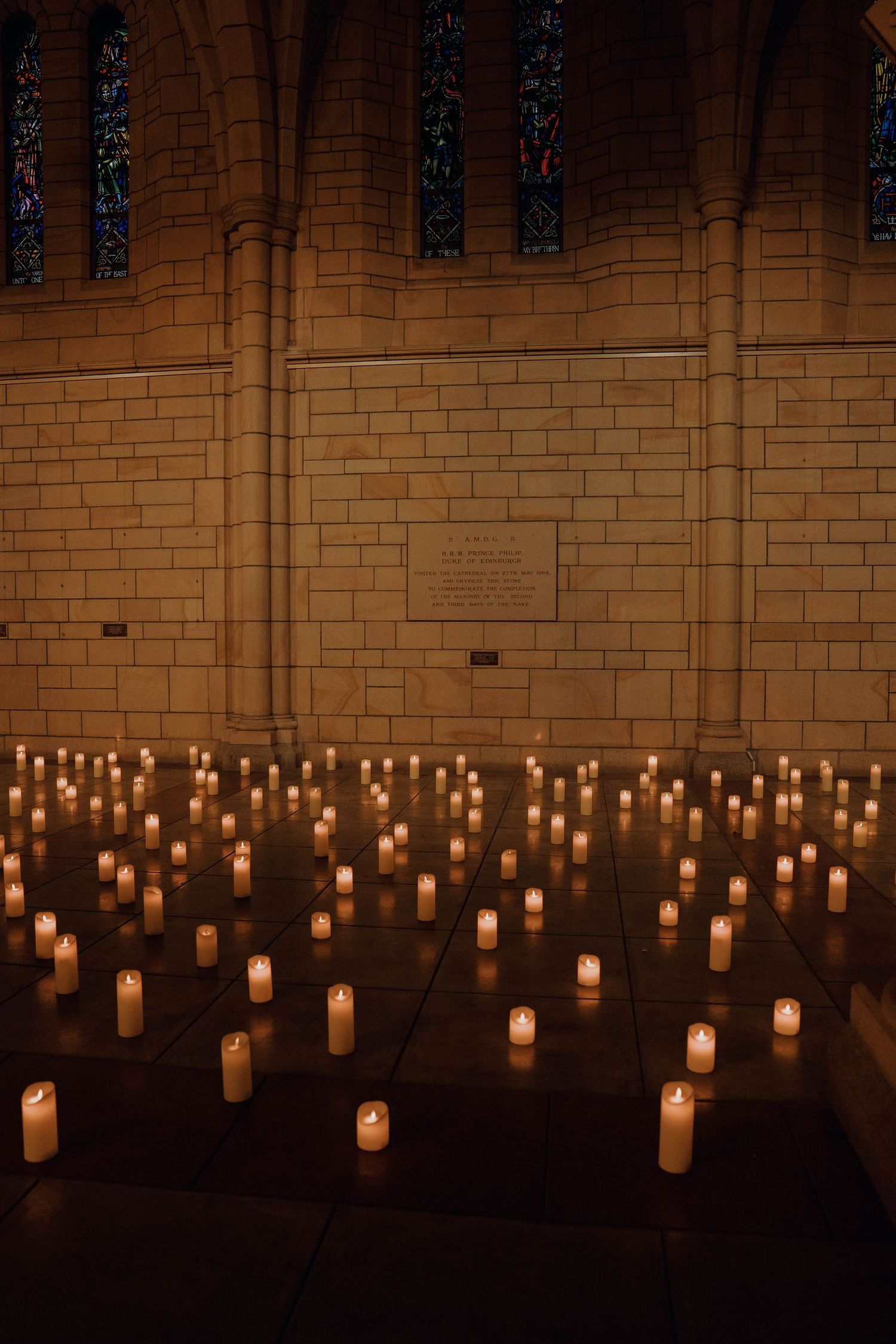 Multiple glowing candles line the floor against a stone wall in a dimly lit church or cathedral interior.