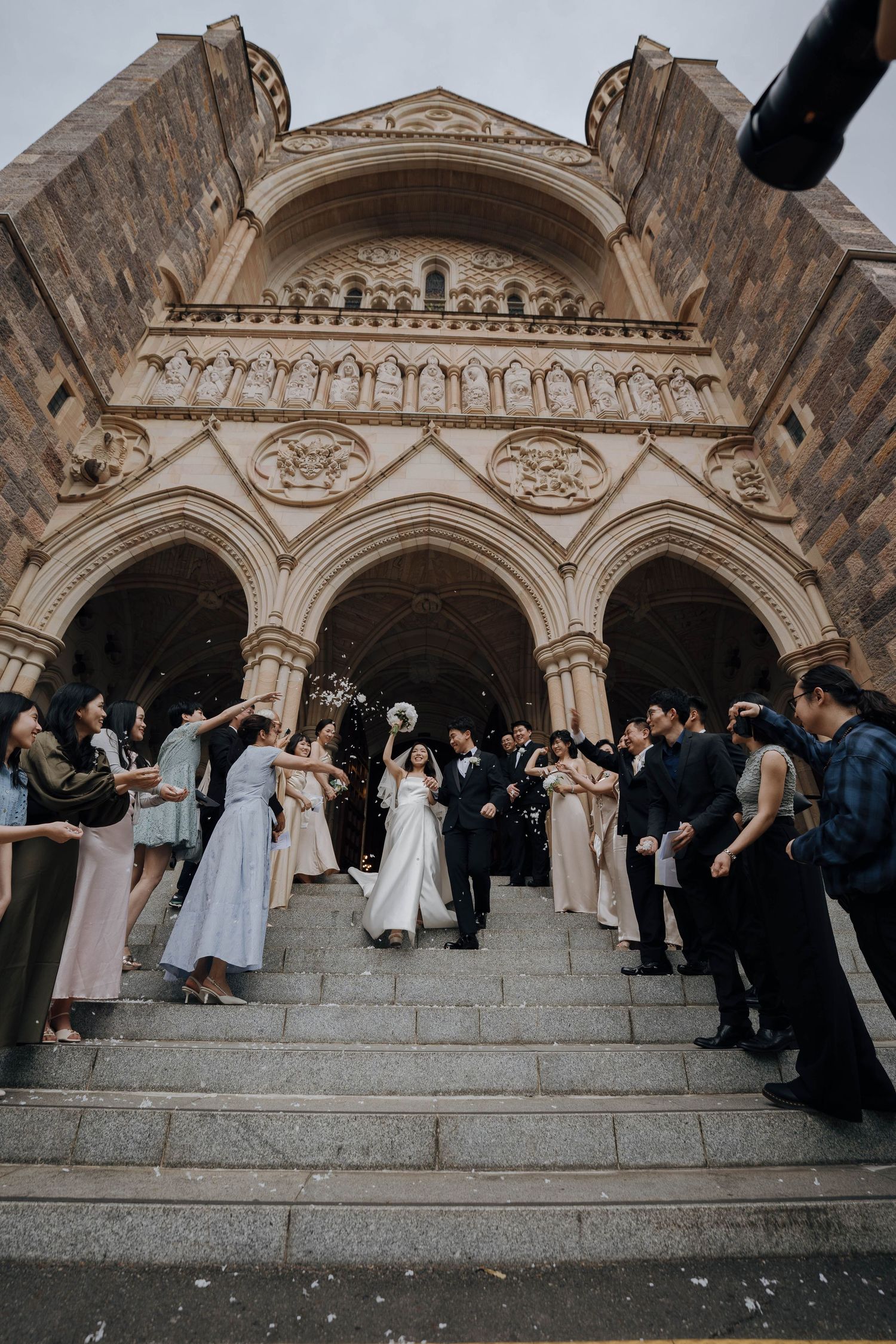 Wedding party stands on stone steps outside ornate church entrance during outdoor ceremony celebration.