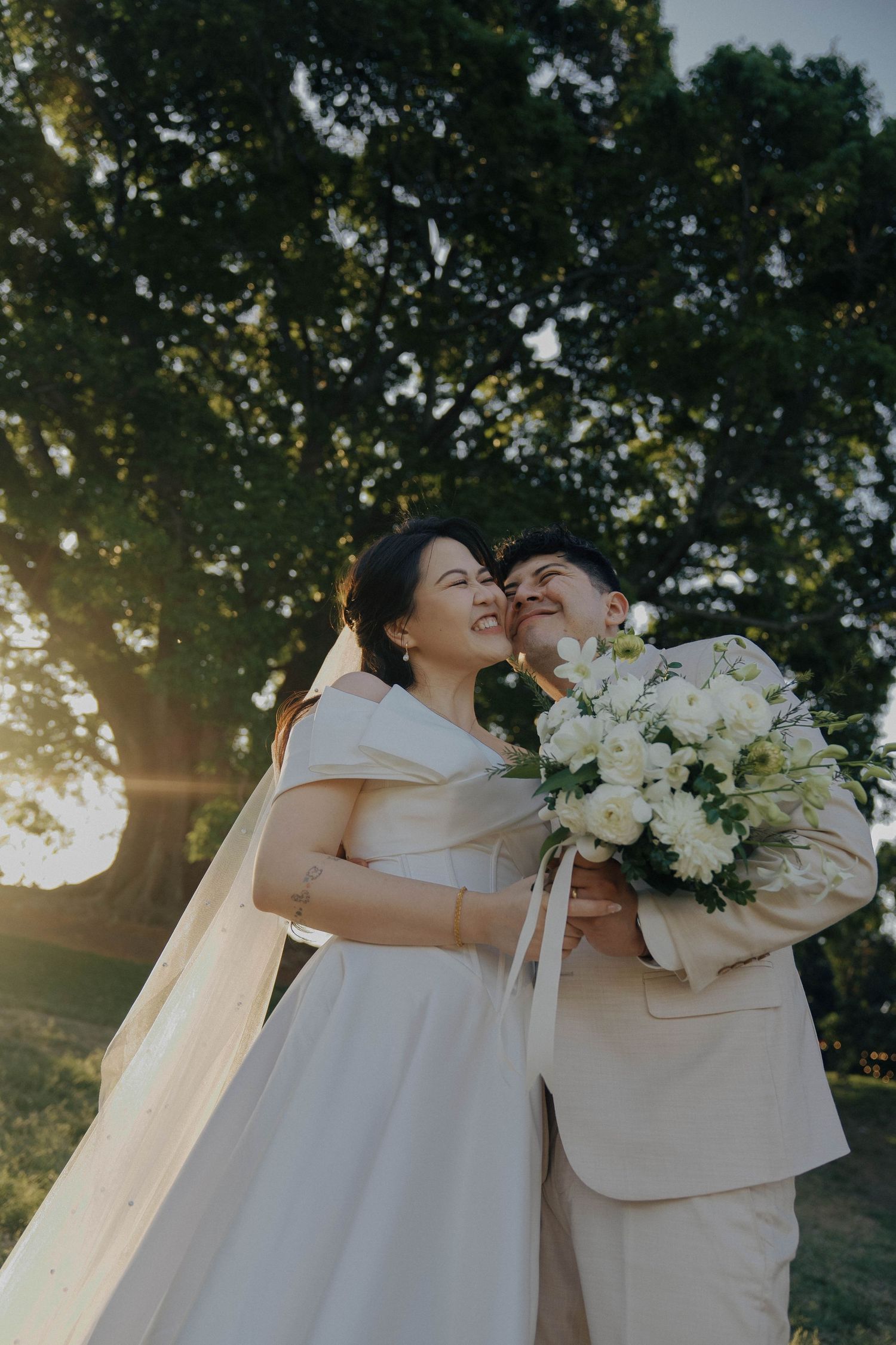 A romantic wedding couple embraces outdoors in natural sunlight with white floral bouquet.