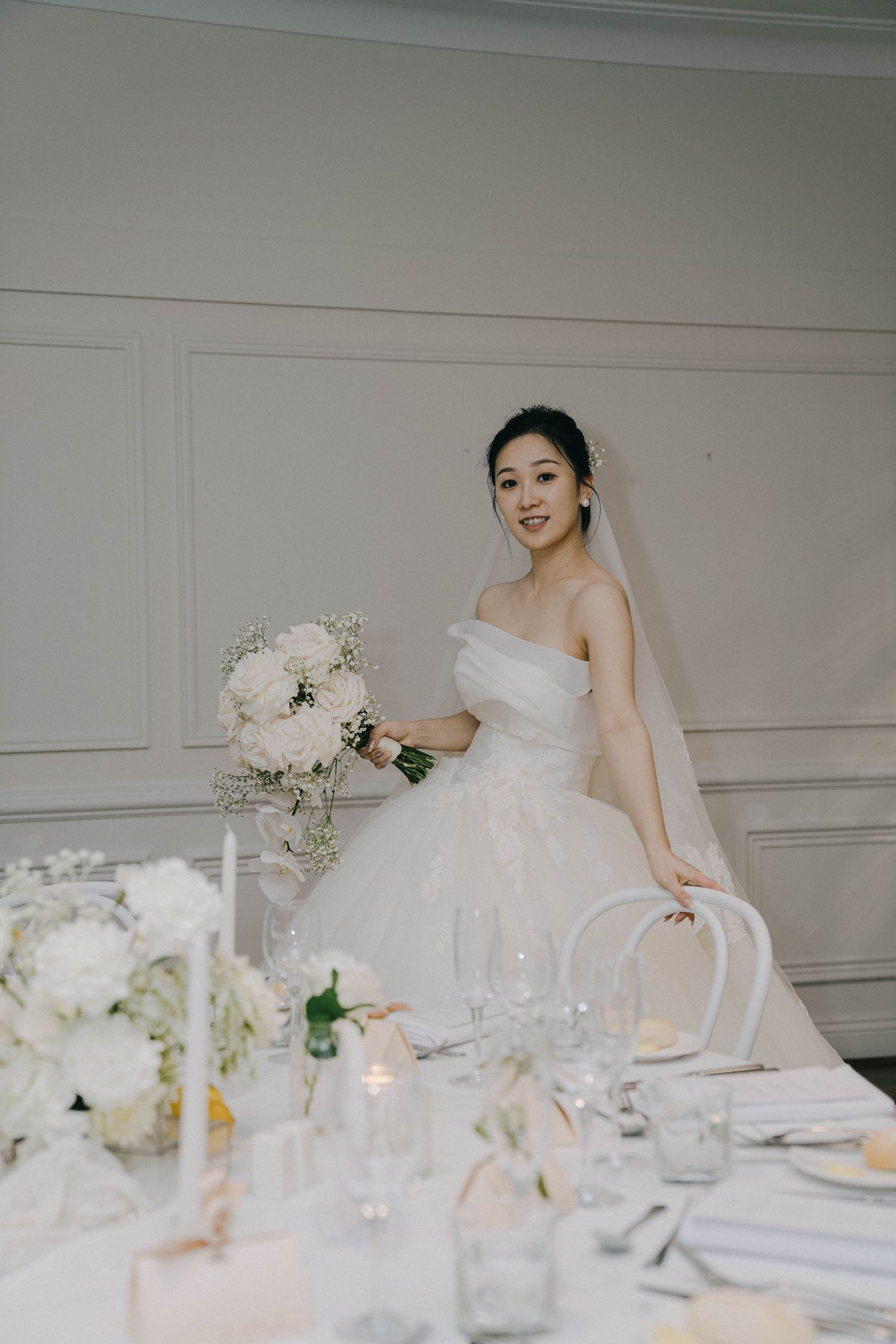 Elegant white wedding reception table setting with floral centerpieces and crystal glassware in a bright room.