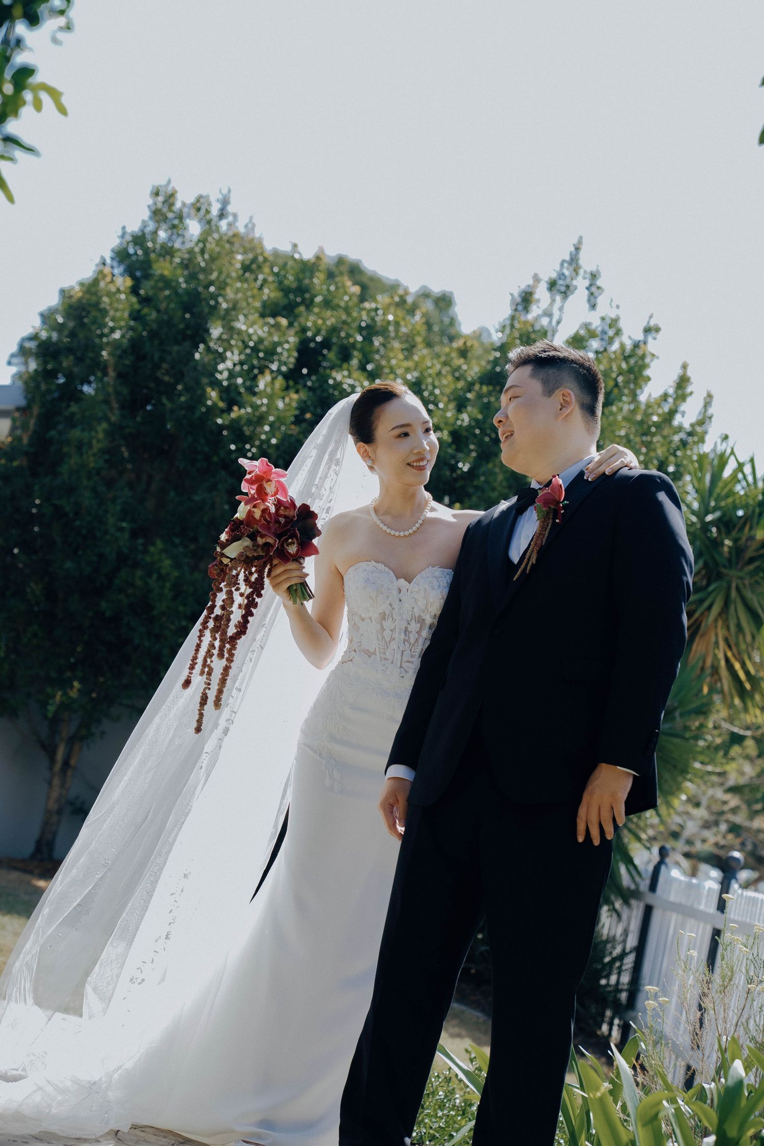 A couple in wedding attire share a romantic moment outdoors on their special day with flowing white veil and greenery behind them.