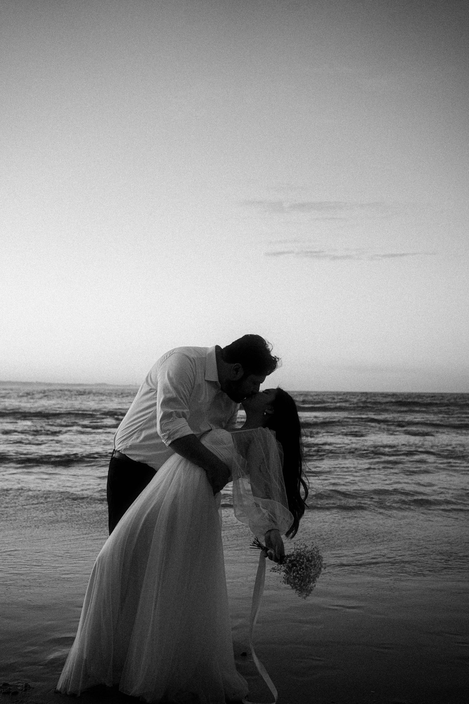 A romantic black and white sequence of a couple in wedding attire sharing an intimate moment by the ocean at sunset.