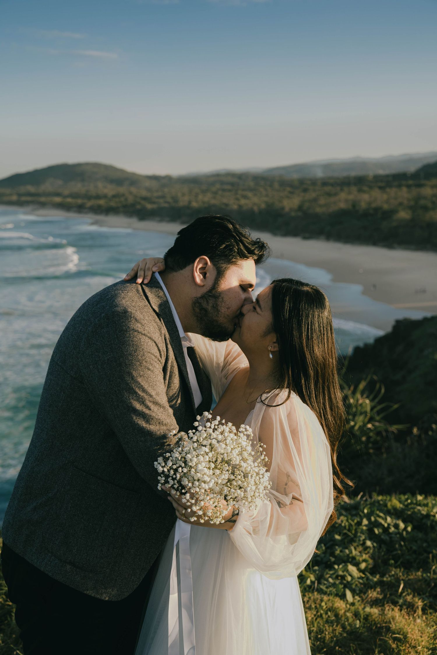 Romantic coastal wedding scene with couple sharing a kiss while holding baby's breath bouquet on cliff overlooking ocean beach.