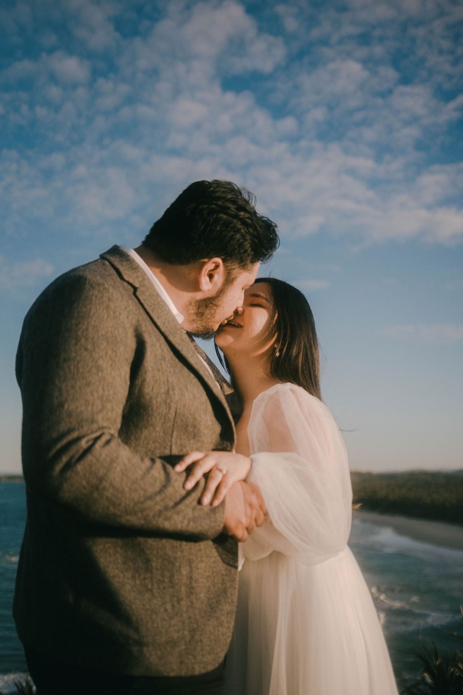 A romantic couple in wedding attire share intimate moments by the ocean under cloudy skies.
