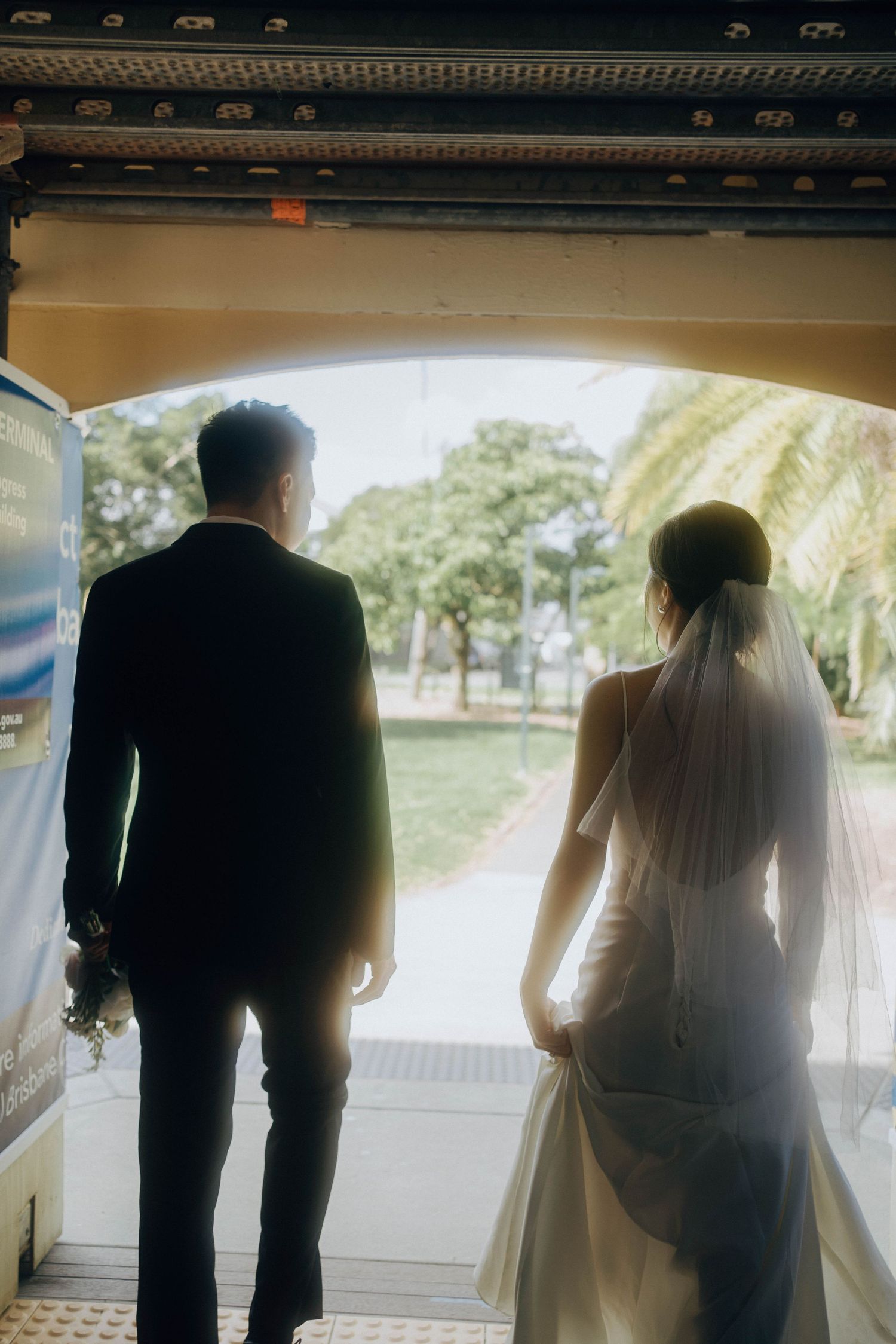 A couple in wedding attire stands silhouetted in an archway overlooking a sunny garden landscape.