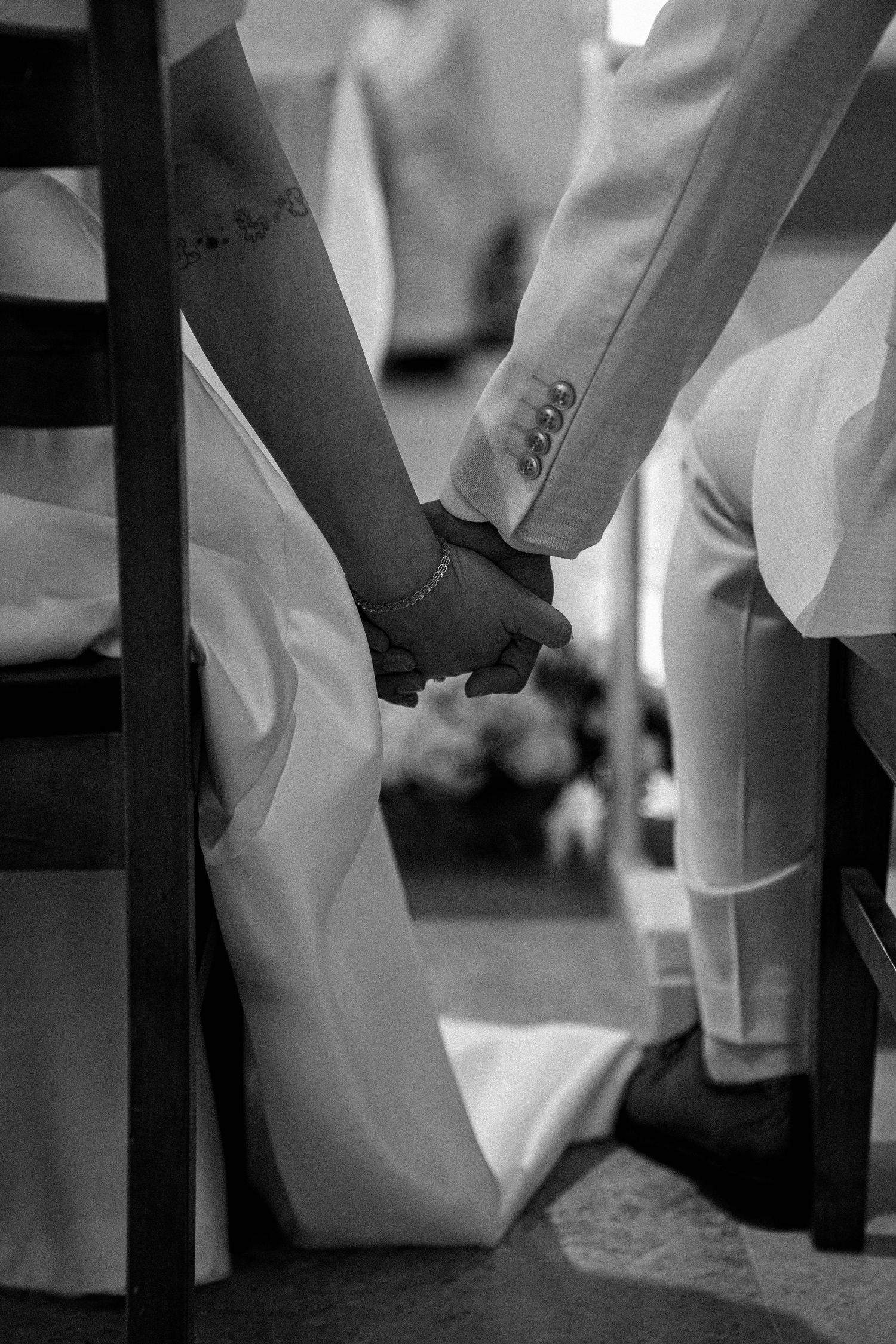 A black and white close-up of two hands holding during a wedding ceremony, with formal attire visible.