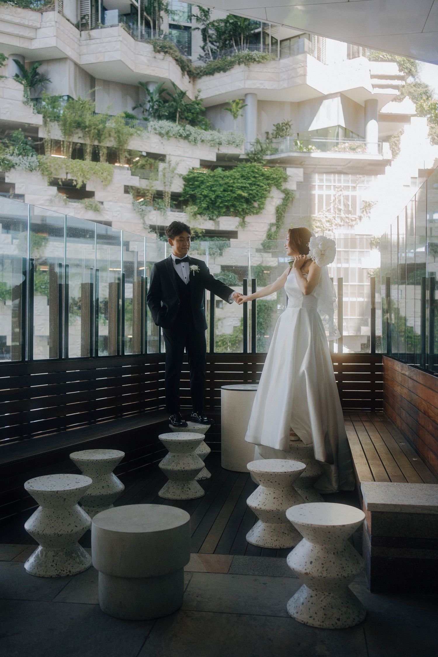 A couple in formal wedding attire stands on a modern balcony with round concrete tables and glass railings.