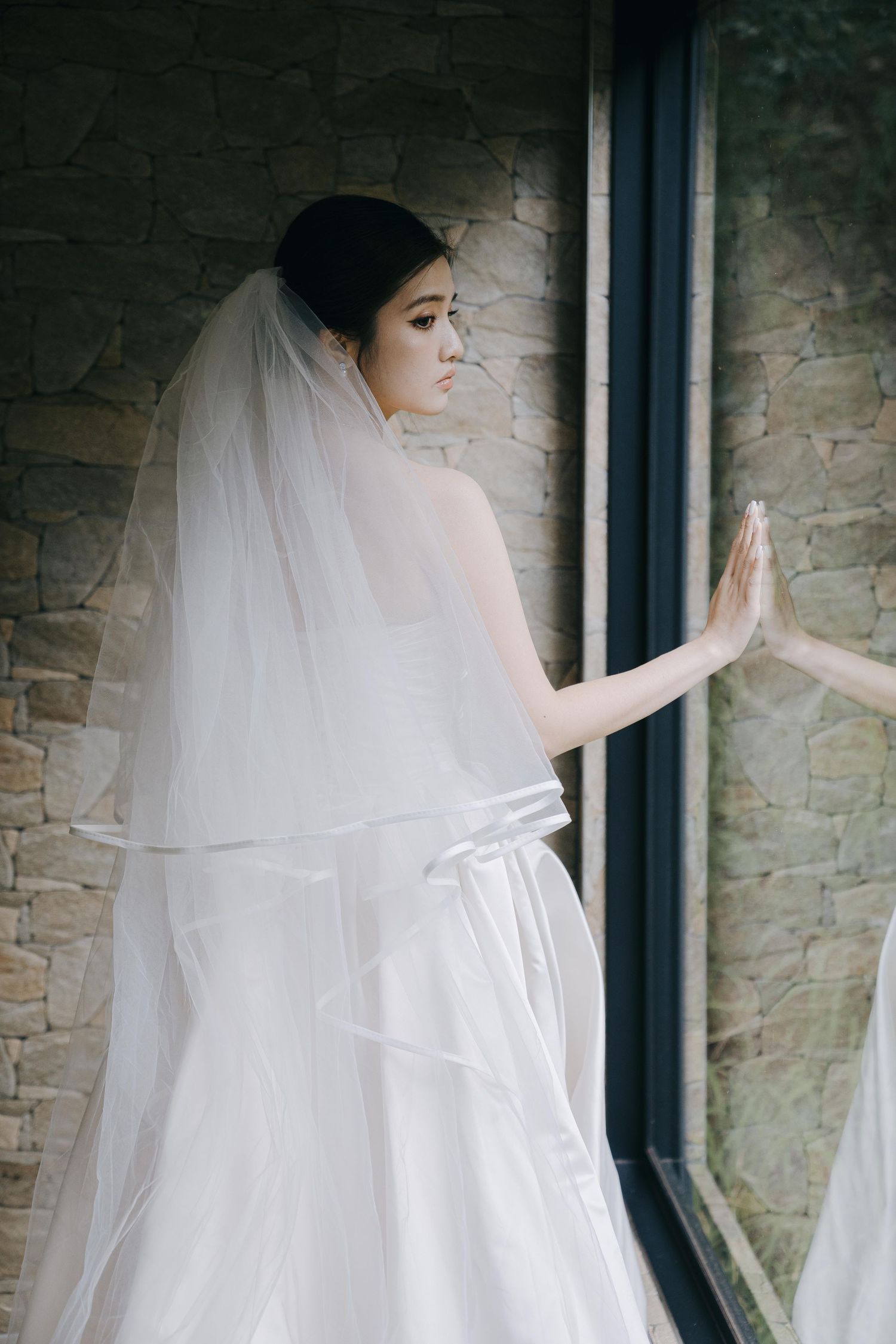A bride in a flowing white gown and veil stands at a window, with her hand pressed against the glass.