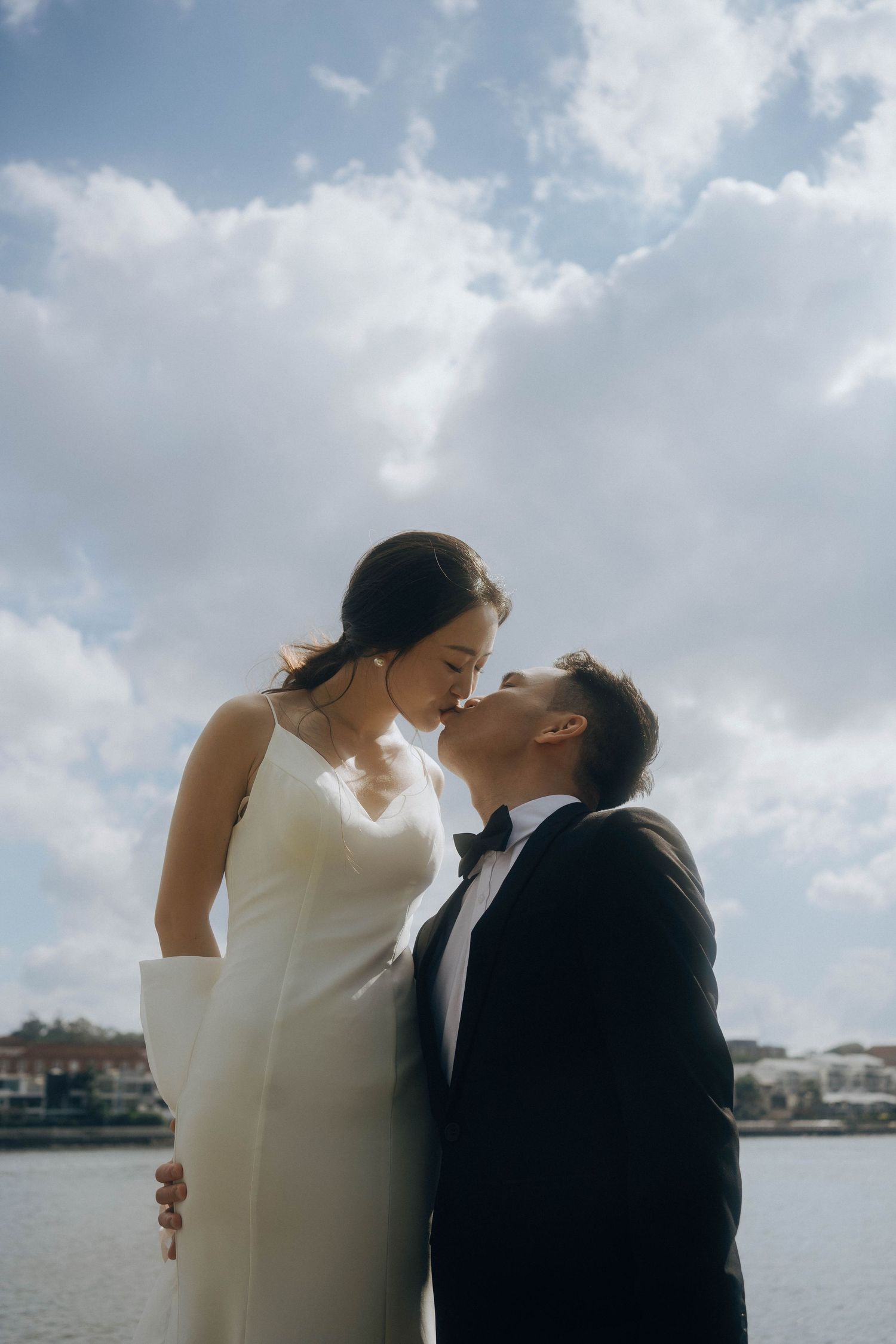 A romantic couple in wedding attire share tender moments against a cloudy waterfront backdrop.