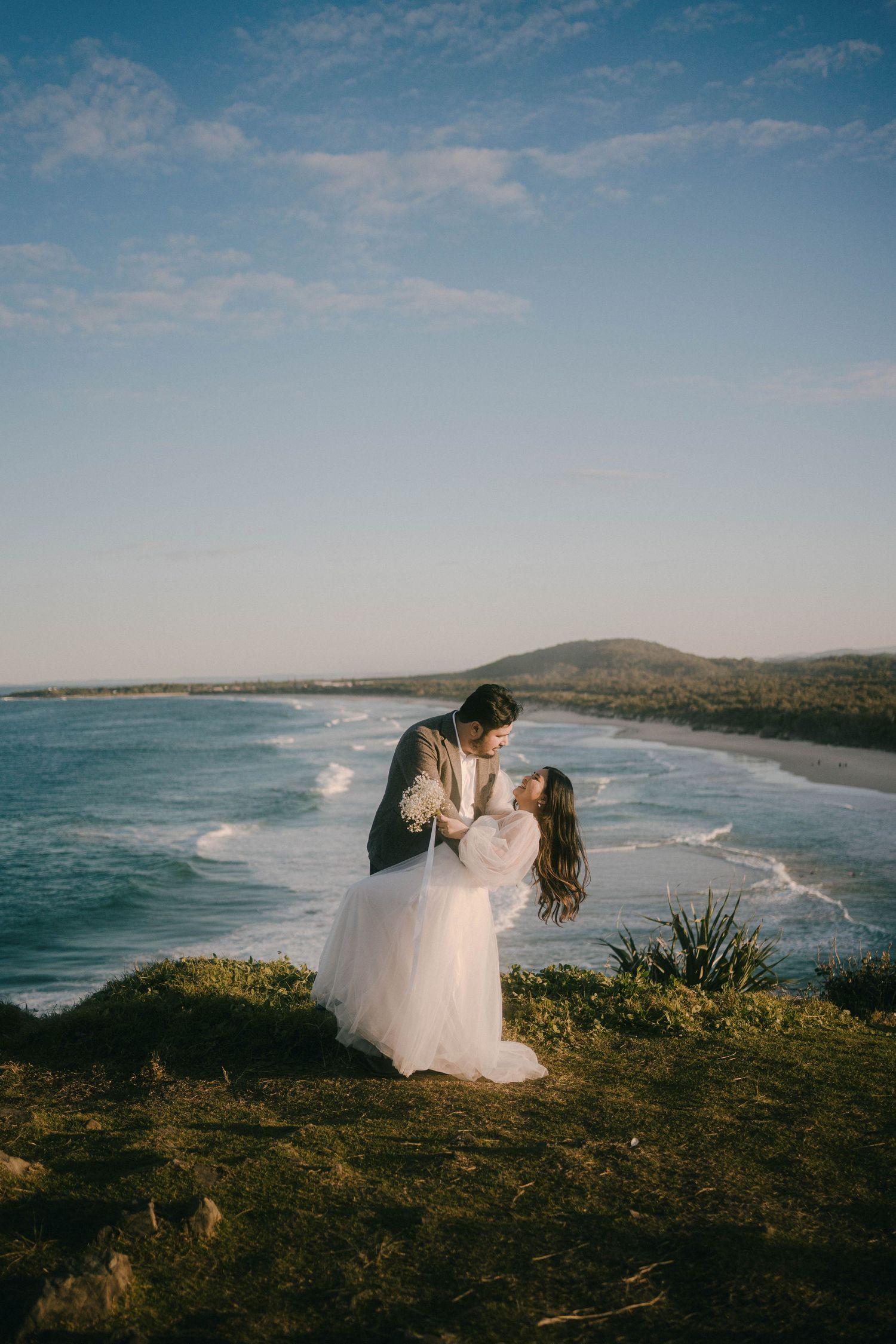 A romantic coastal wedding photoshoot at sunset with dramatic ocean views and cliffside scenery in the background.