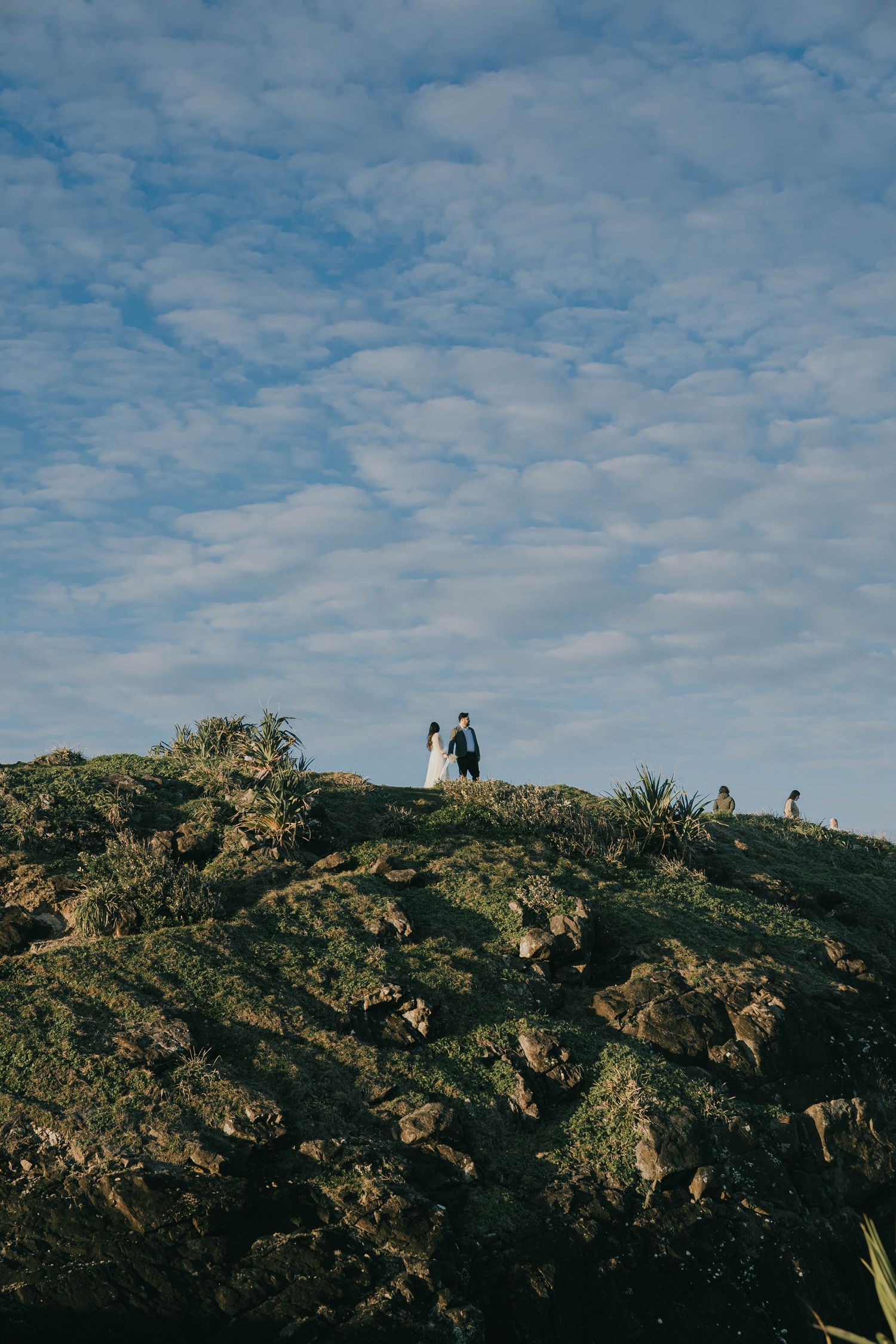 Silhouetted figures stand atop a rocky hill against a bright blue sky with scattered white clouds.