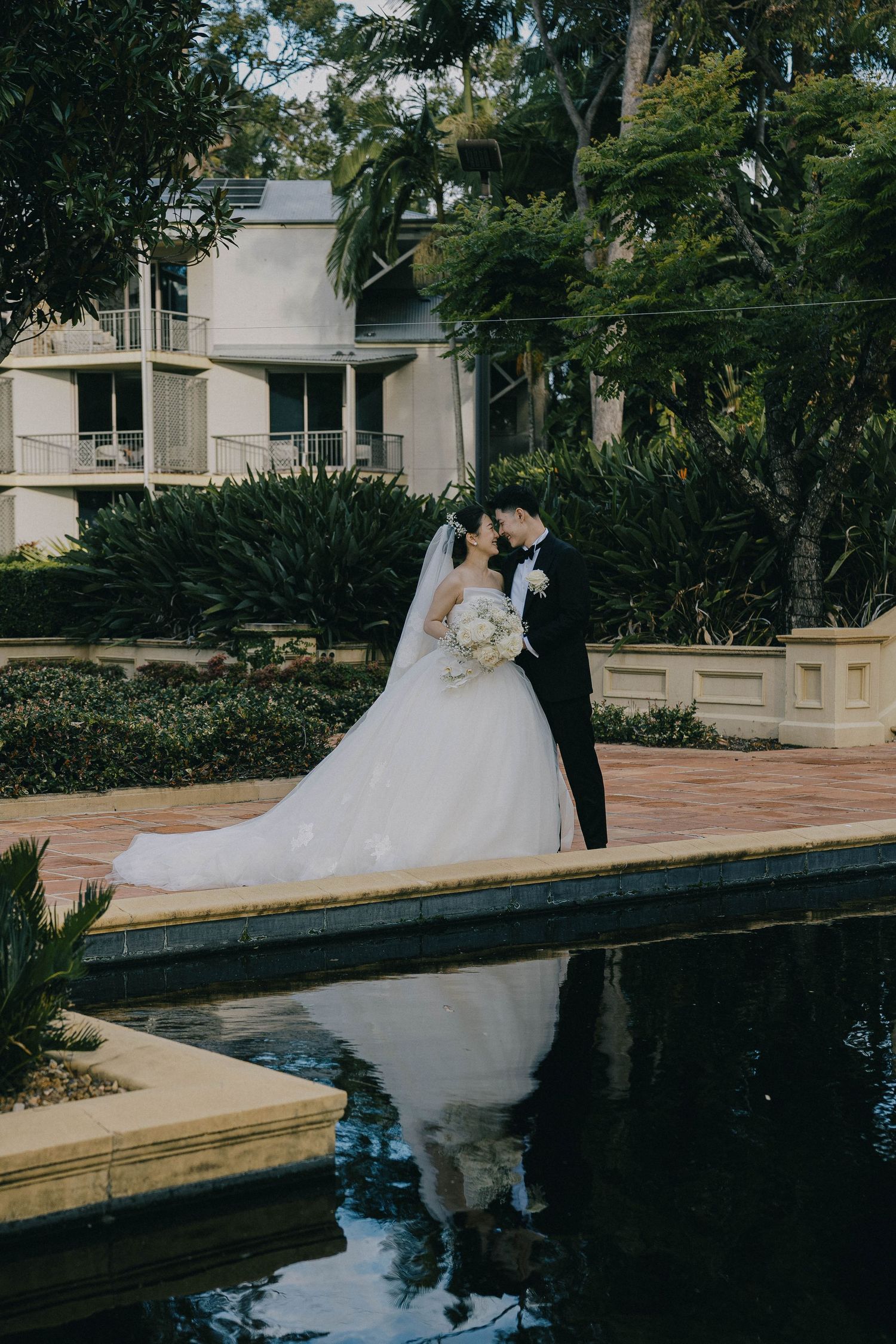 A romantic wedding couple poses together by a reflective pool in an elegant garden setting at dusk.