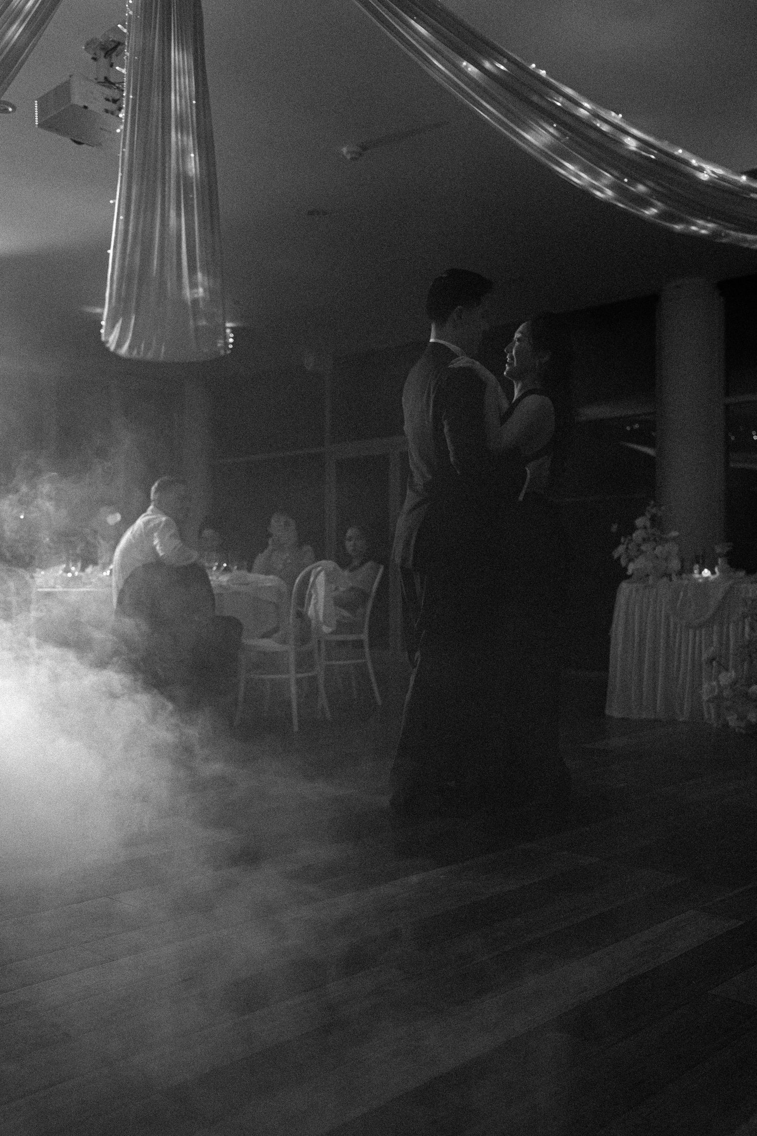 Atmospheric fog or smoke fills a dimly lit indoor space with silhouetted furniture and hanging lights creating a moody ambiance.