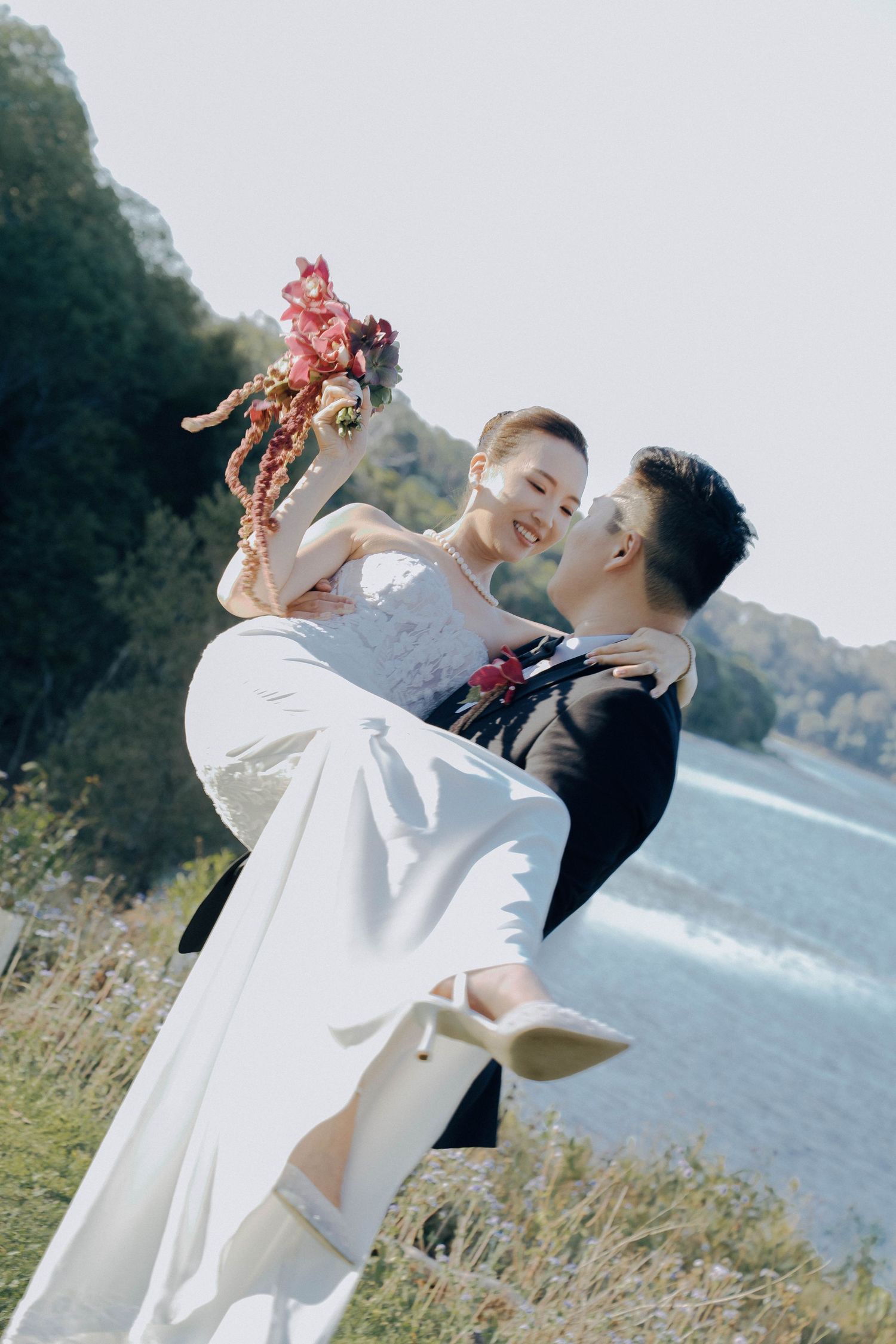 A couple in wedding attire share a romantic moment by a waterfront, with the groom lifting the bride in a flowing white dress.