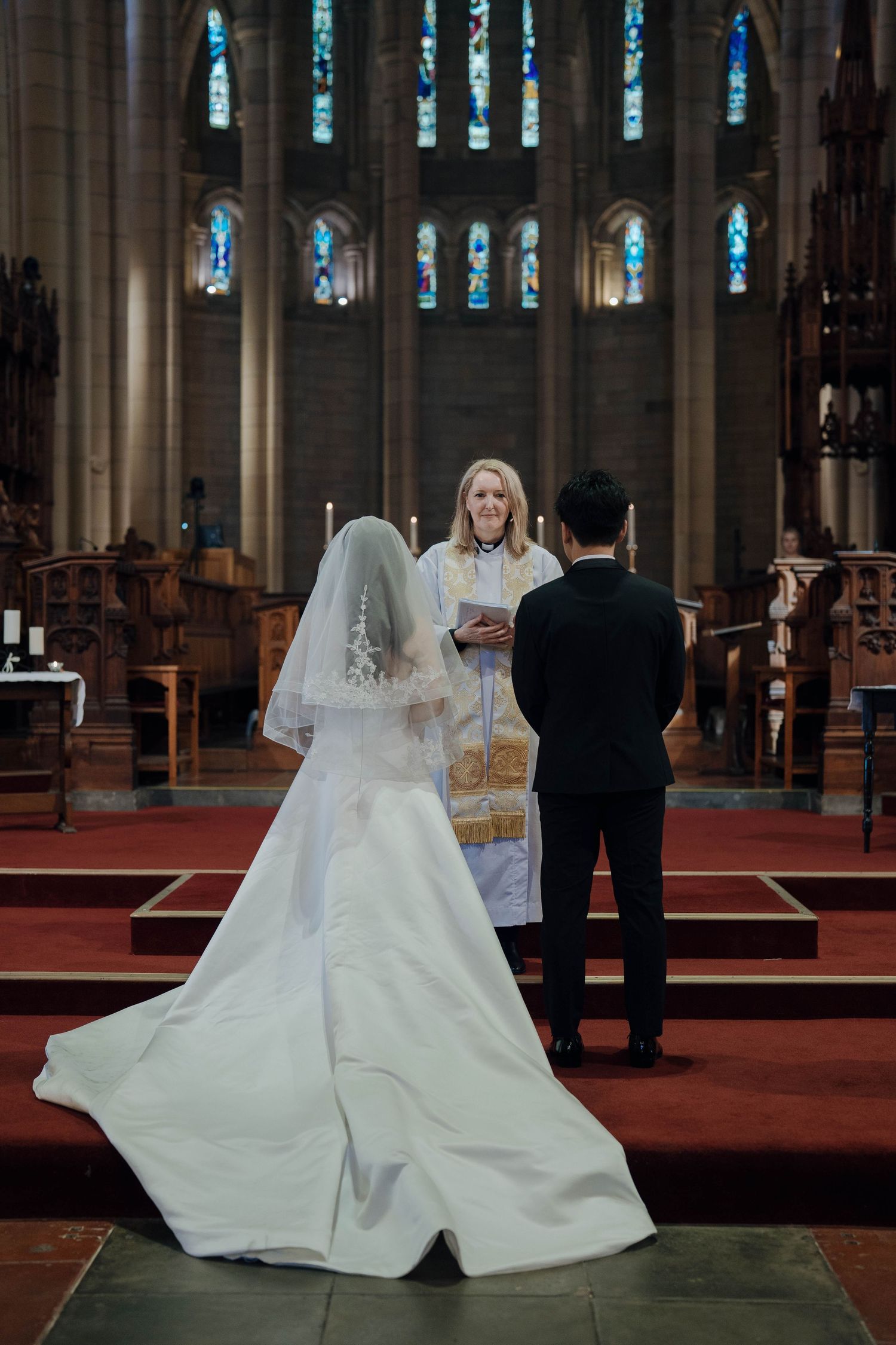 A wedding ceremony takes place inside a grand cathedral with stained glass windows and dramatic lighting.