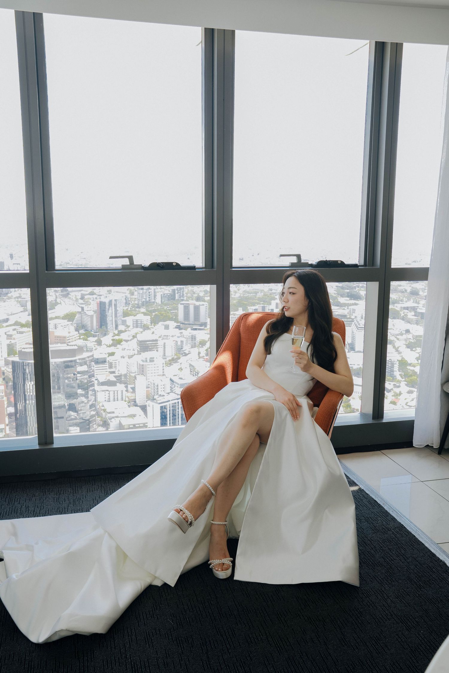 A bride in a white wedding dress reclines elegantly on a dark floor against floor-to-ceiling windows overlooking a city.