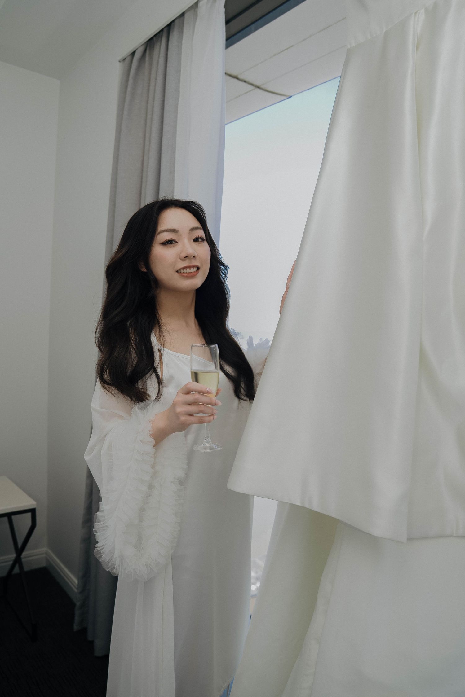 A person in a white fluffy robe holds a champagne glass while standing near white curtains in a bright room.