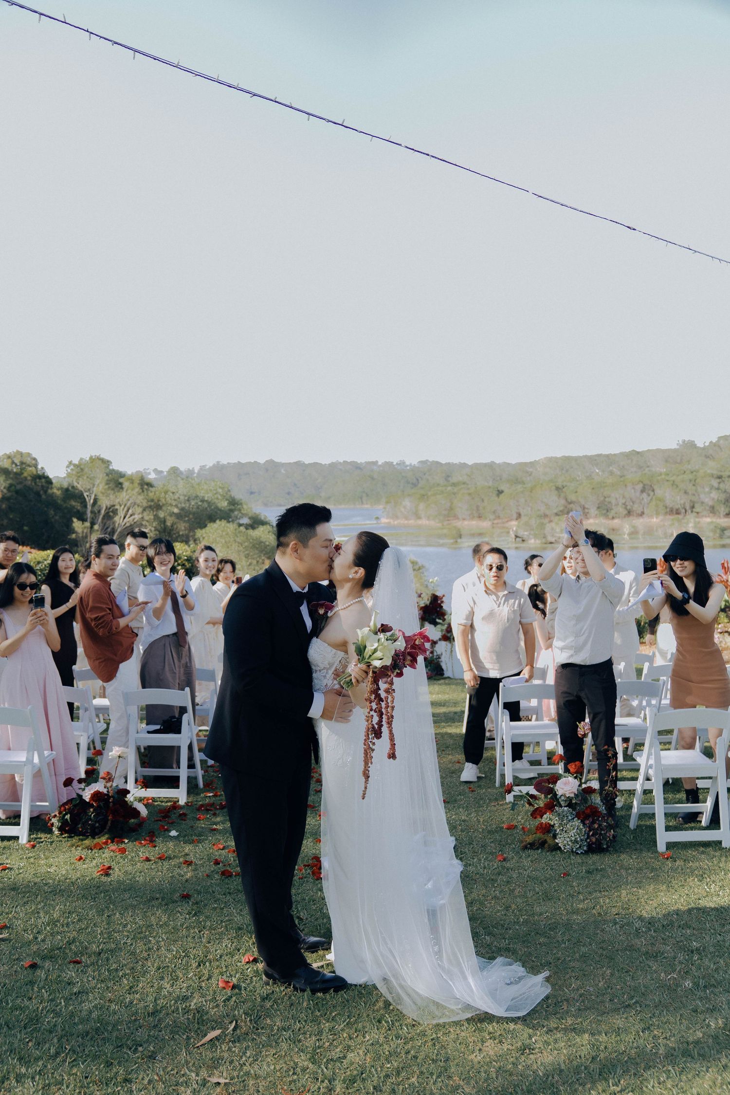 A couple shares a romantic moment during their outdoor wedding ceremony as guests look on from white chairs on a grassy field.
