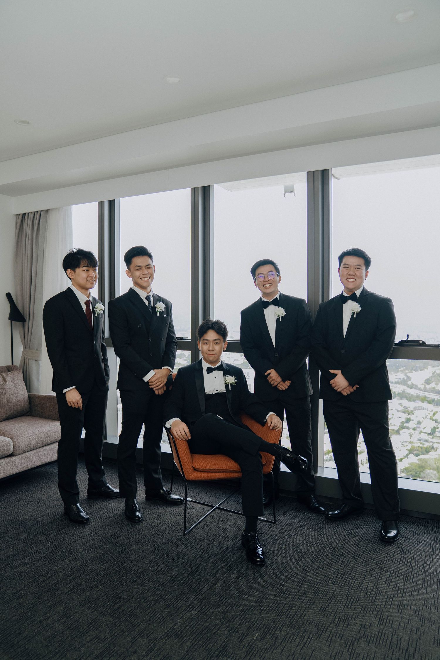 Groomsmen in black tuxedos and bow ties pose playfully in a modern room with floor-to-ceiling windows.