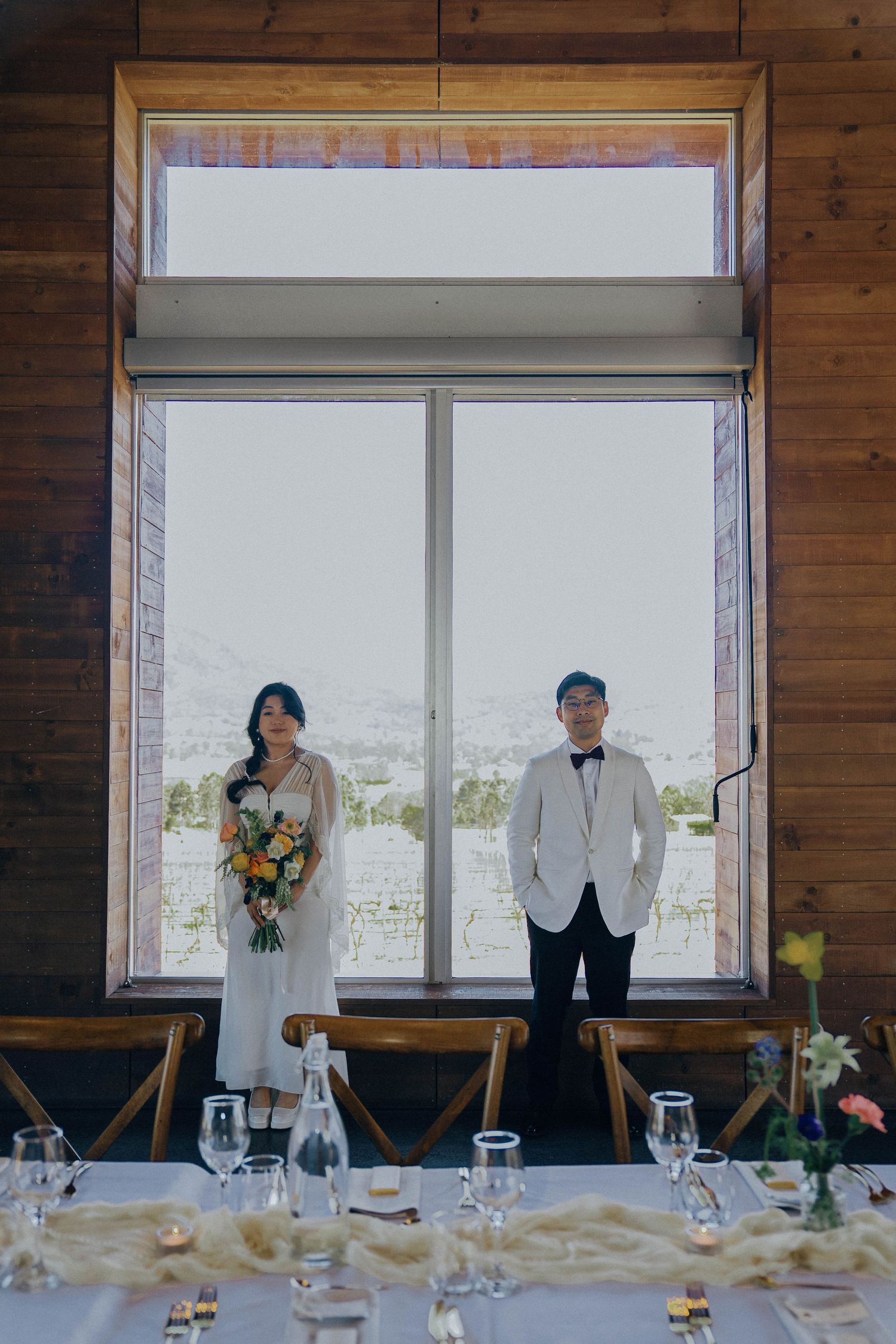 A couple stands at a formal dining table setup with glassware and decor against large windows in a rustic wooden room.