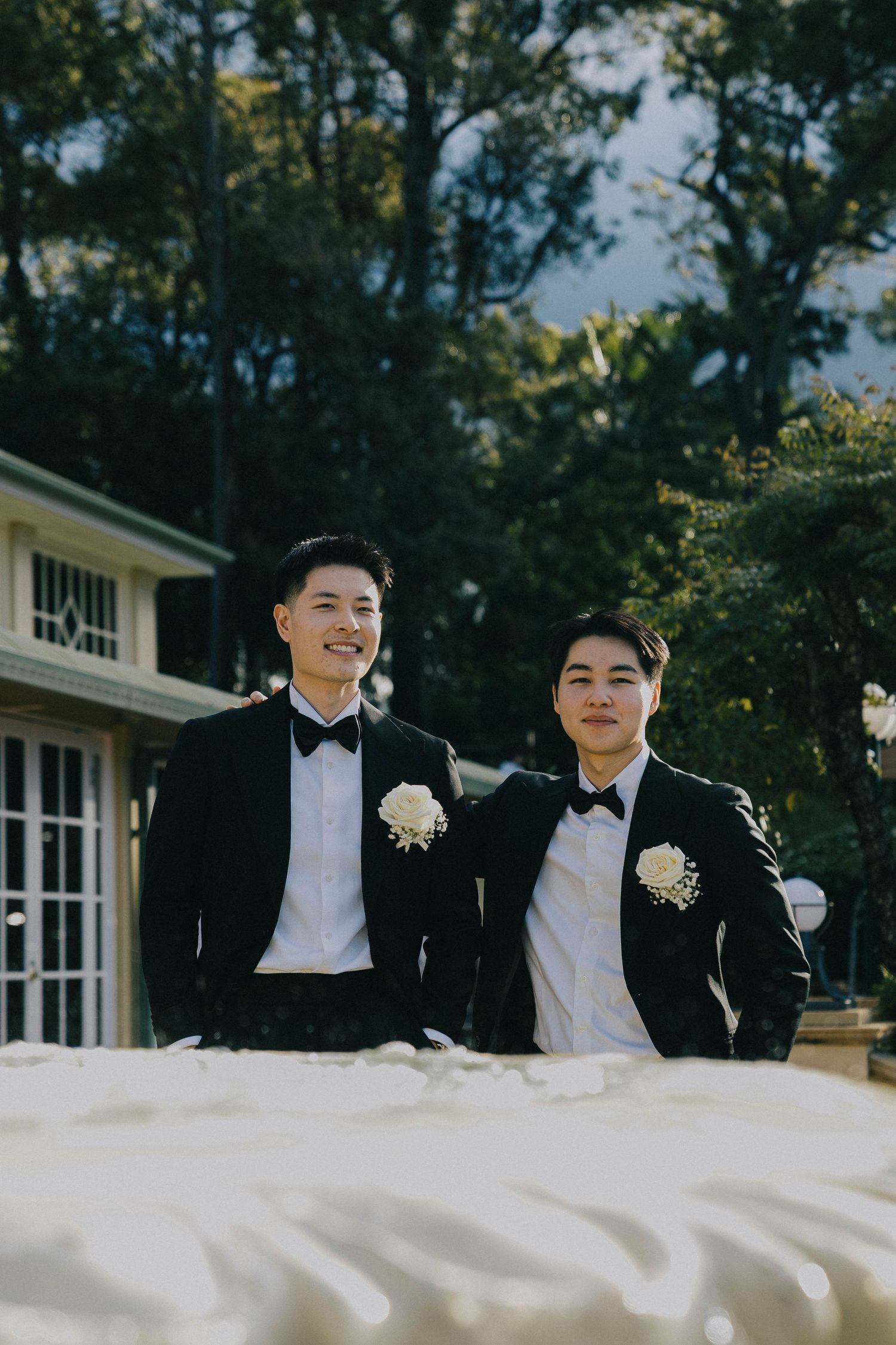 Two groomsmen in black tuxedos and bow ties share a candid moment outdoors at a wedding venue.