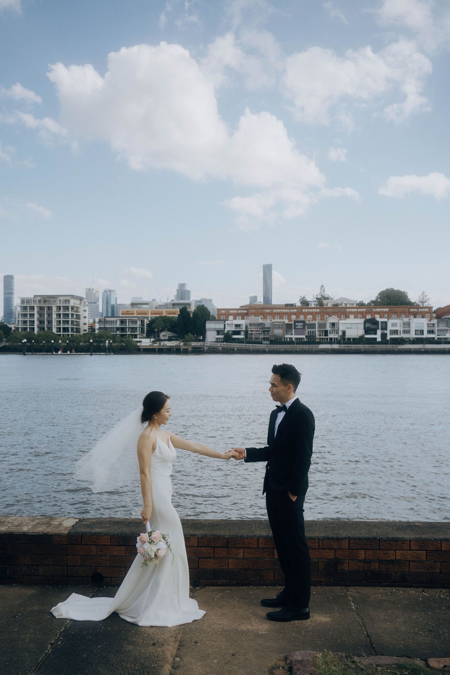 A couple in wedding attire shares romantic moments along a waterfront with a cityscape backdrop.
