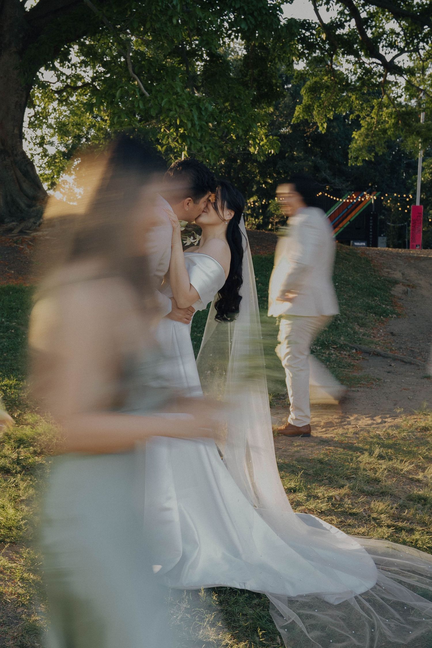 Artistic motion blur wedding photo shows couple in white attire embracing under green trees at outdoor ceremony.