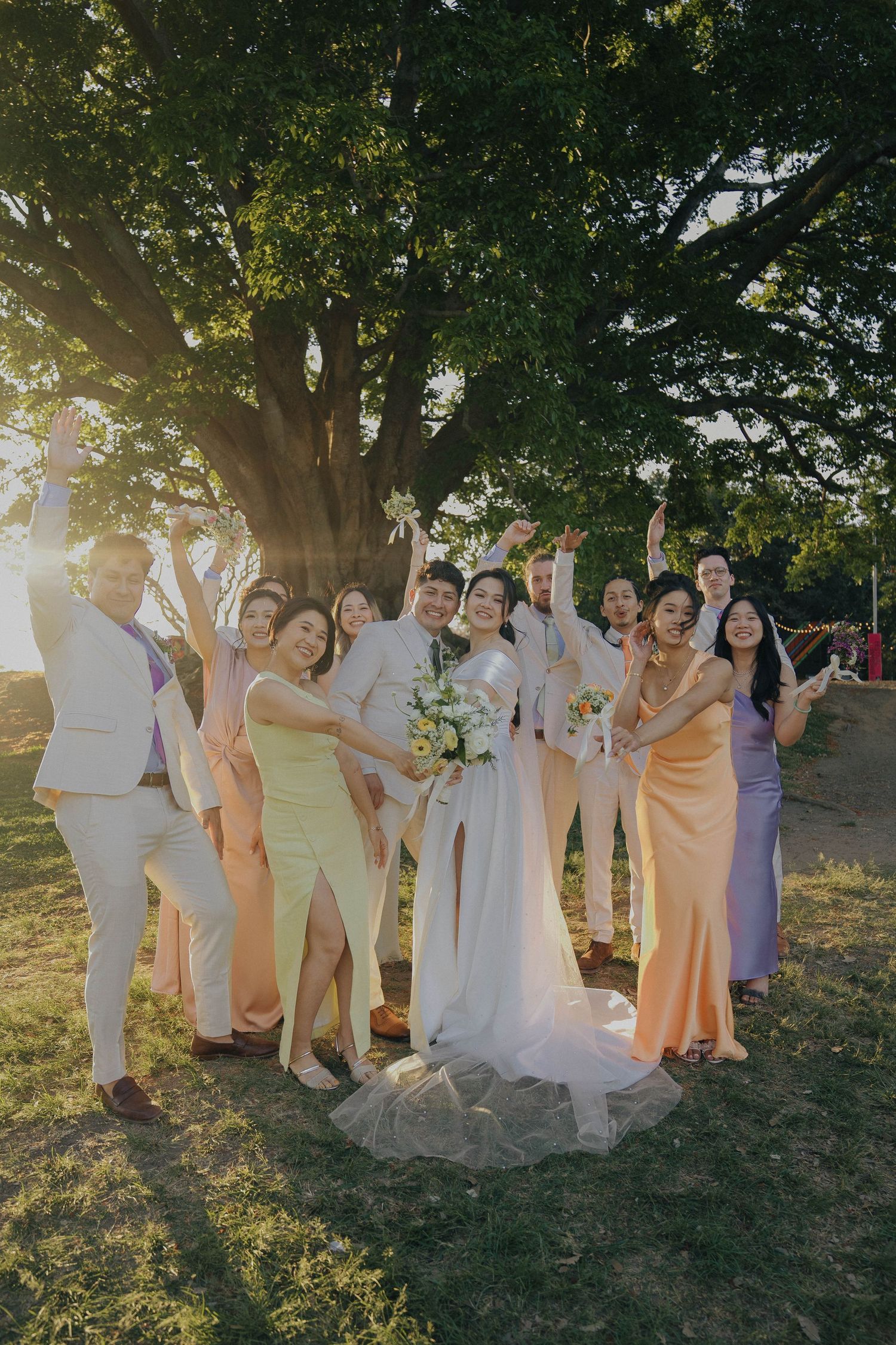 A wedding party in pastel dresses poses together outdoors during golden hour sunlight filtering through trees.
