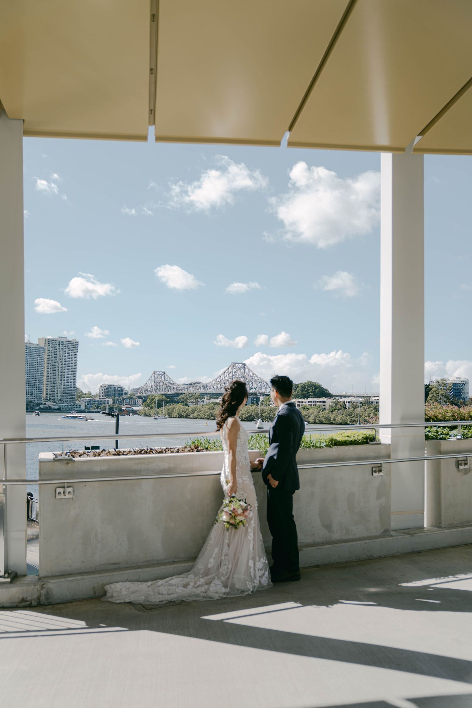 A couple poses for wedding photos on a bright outdoor terrace overlooking a cityscape with white columns and walls.