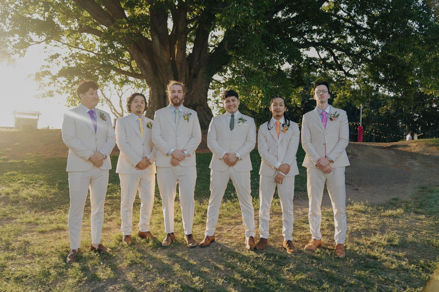 Wedding party in white attire poses together on grassy hillside during golden hour sunset.