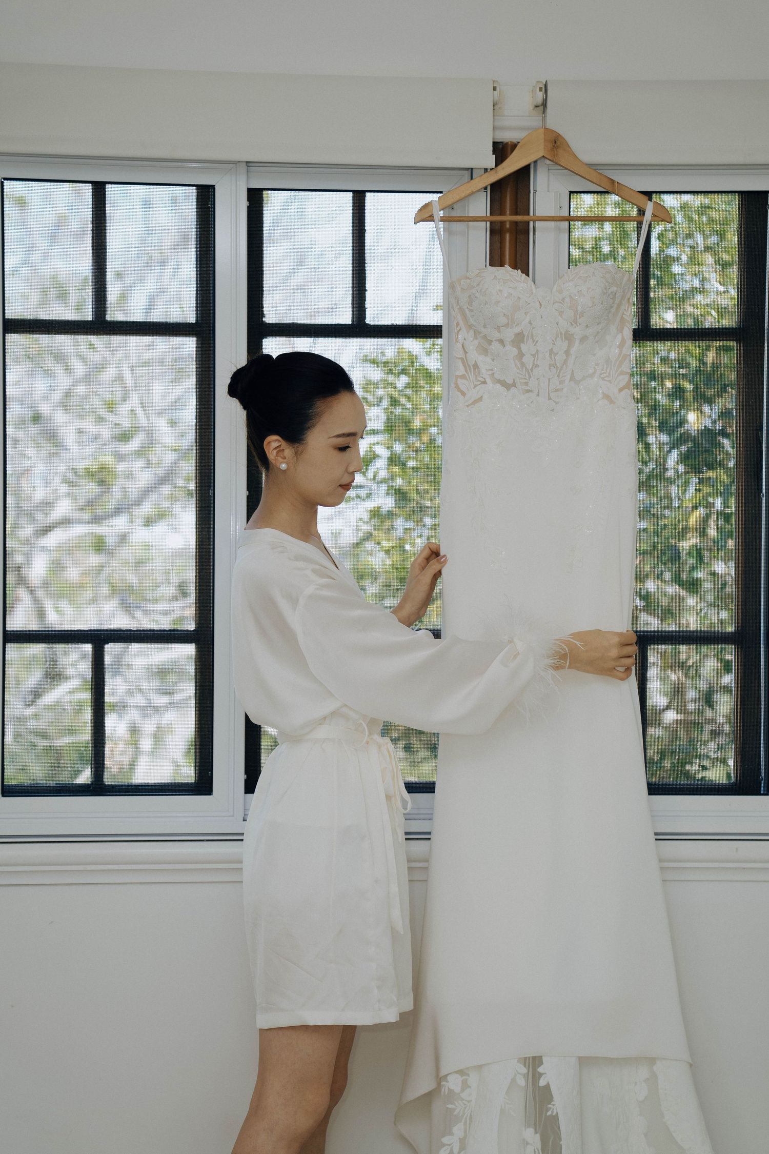 A wedding dress hangs on display in a bright sunlit room while someone in white examines it.