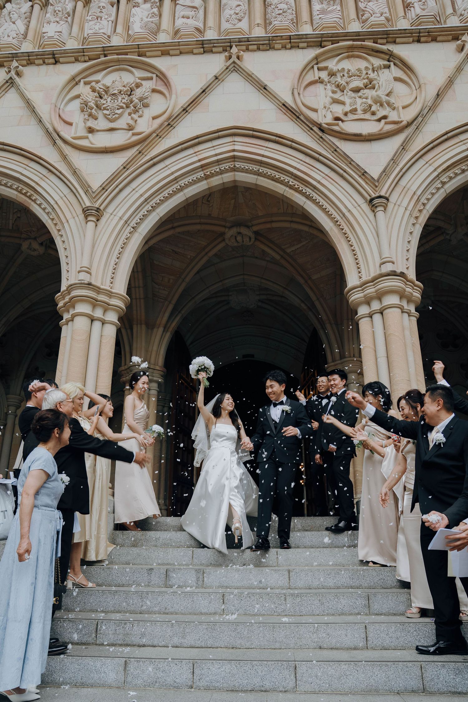 Wedding party celebrates with bubbles on grand stone steps under ornate arched colonnade at historic venue.