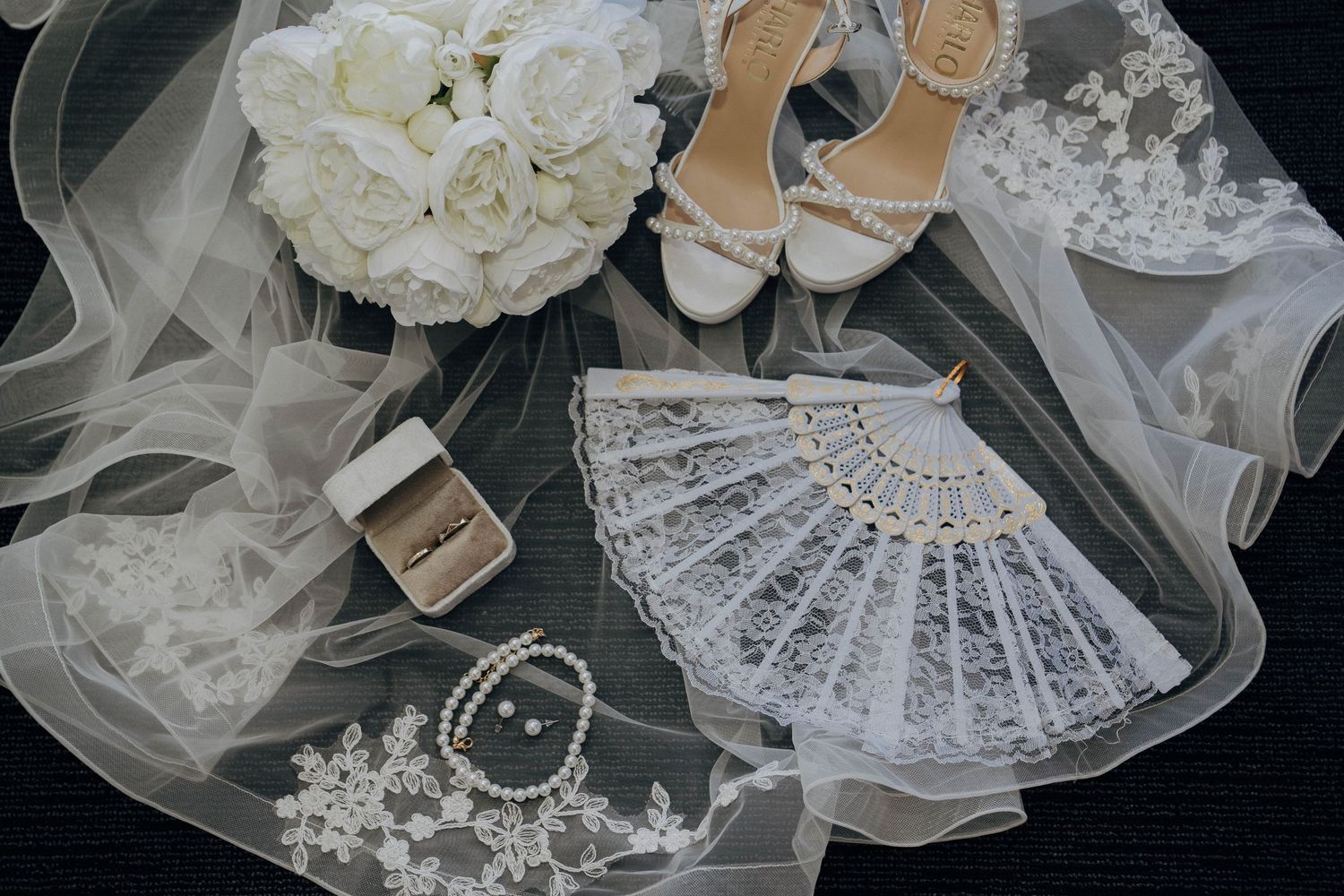 Wedding accessories including white roses, lace details, pearls, and delicate tulle arranged on dark surface.