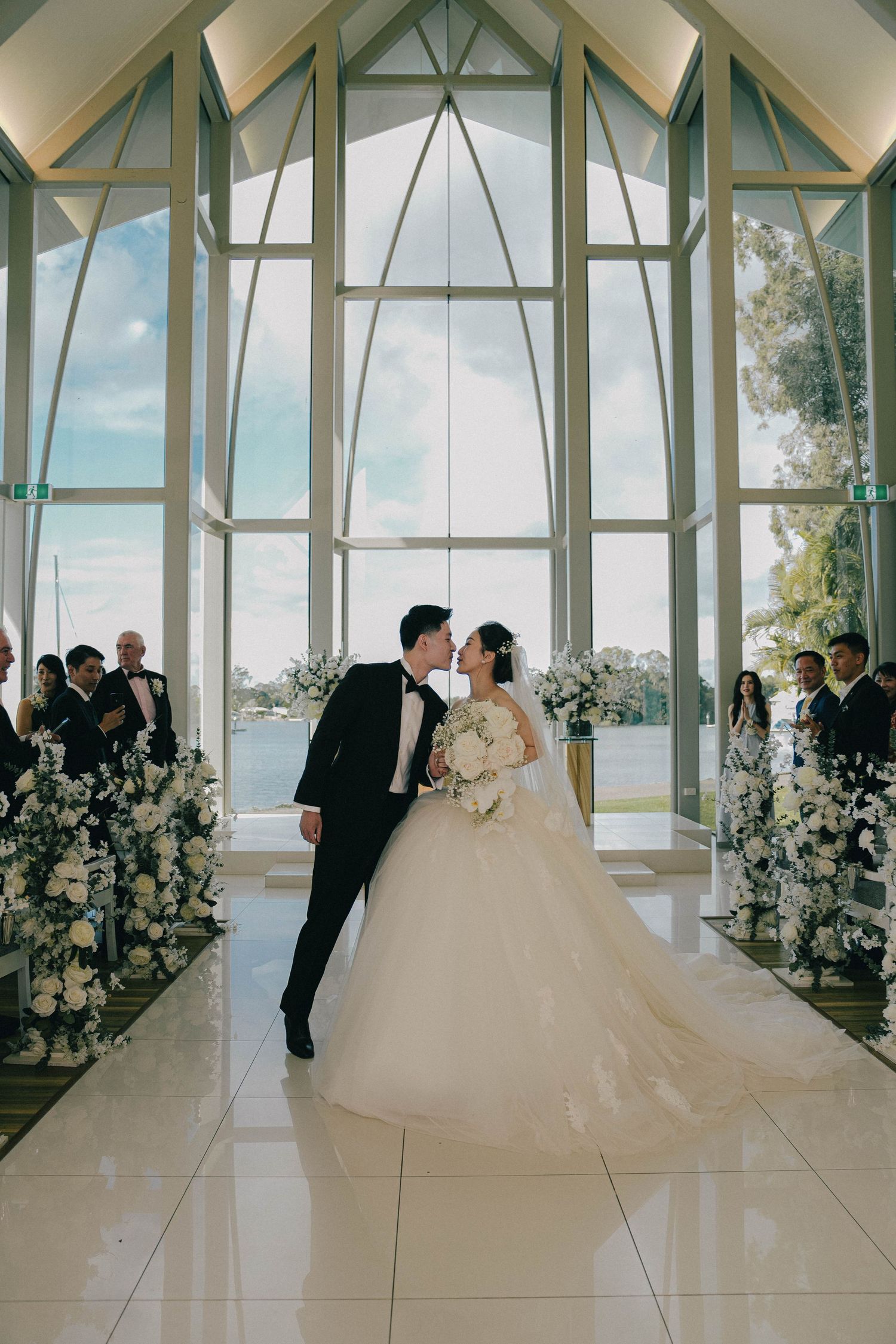 A sequence of five wedding photos showing a couple in a modern glass chapel with white floral arrangements.