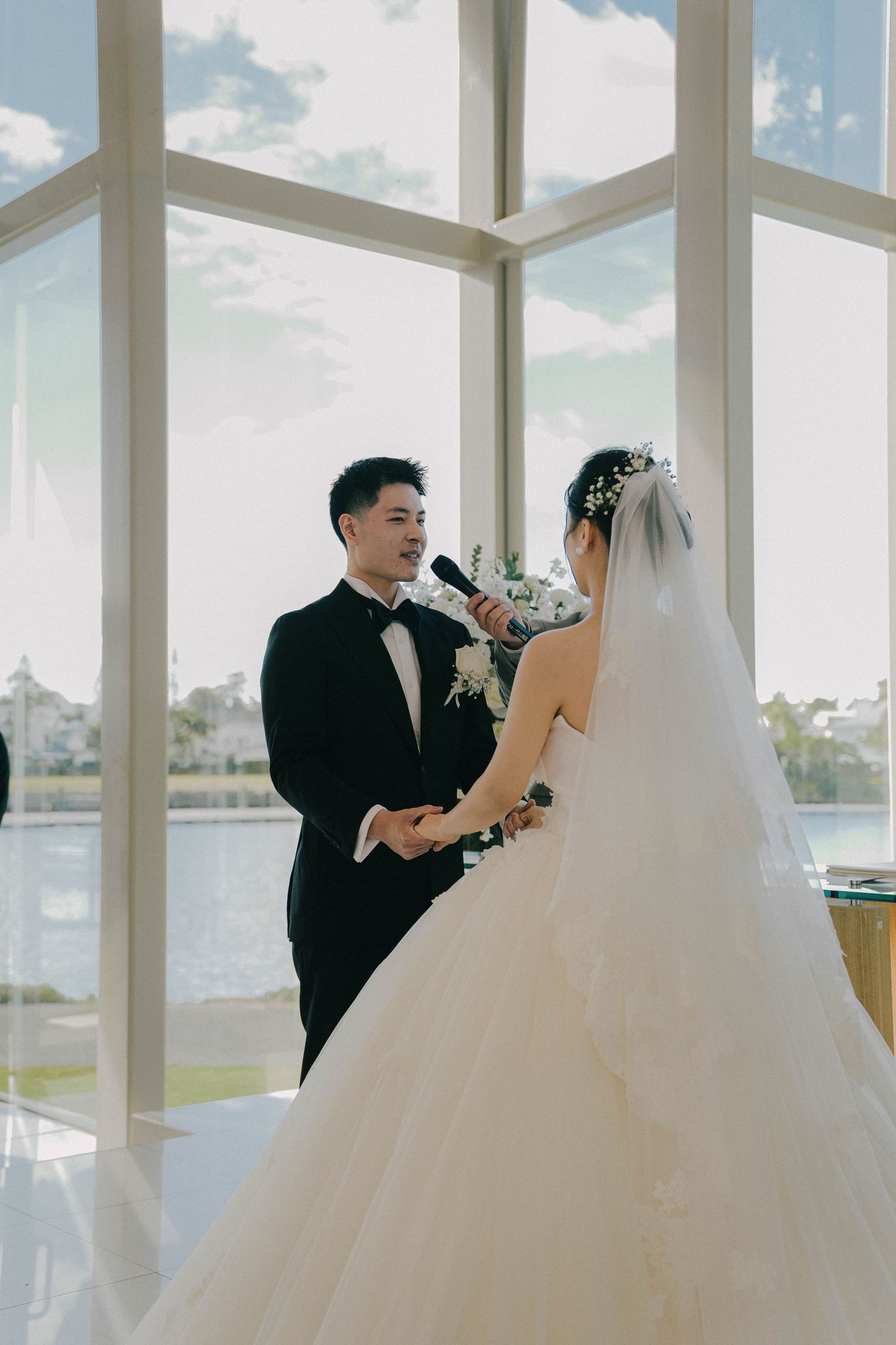 A wedding couple exchanges vows in an elegant glass chapel with waterfront views and natural light streaming in.