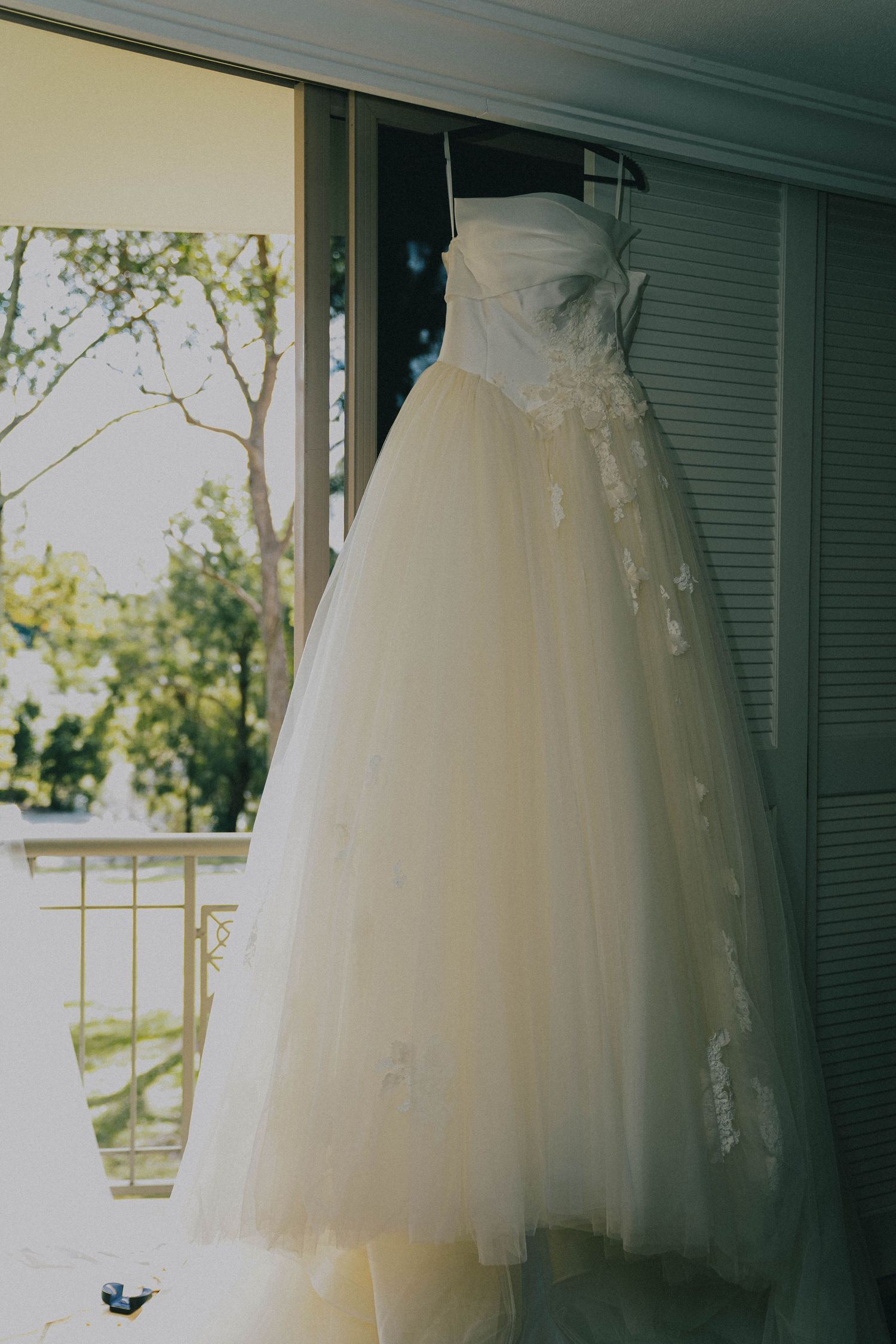 A white tulle wedding dress with lace details hangs elegantly by a window with trees visible in the background.