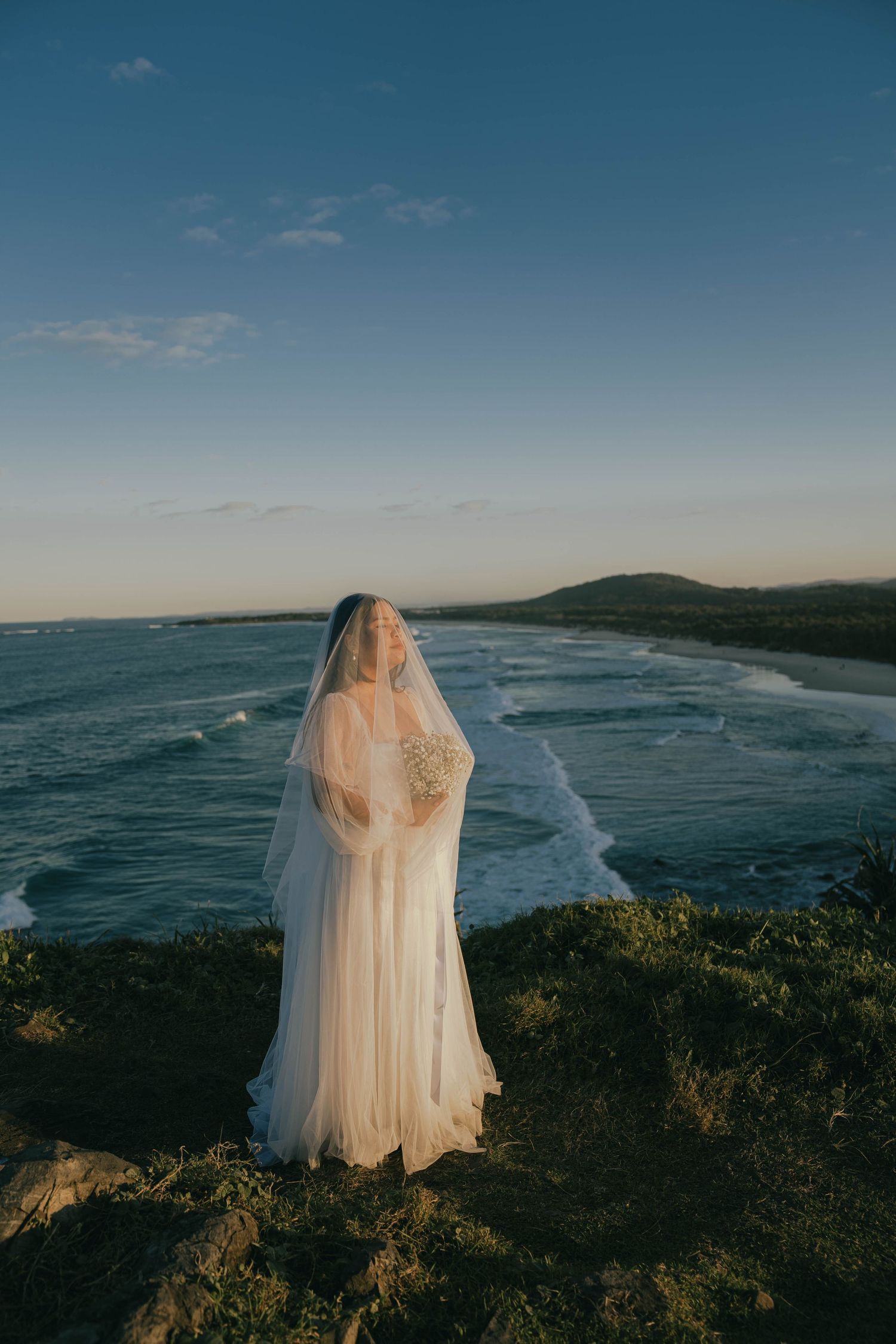 A white wedding dress silhouetted against a dramatic coastal landscape with crashing waves and cliffs at sunset.