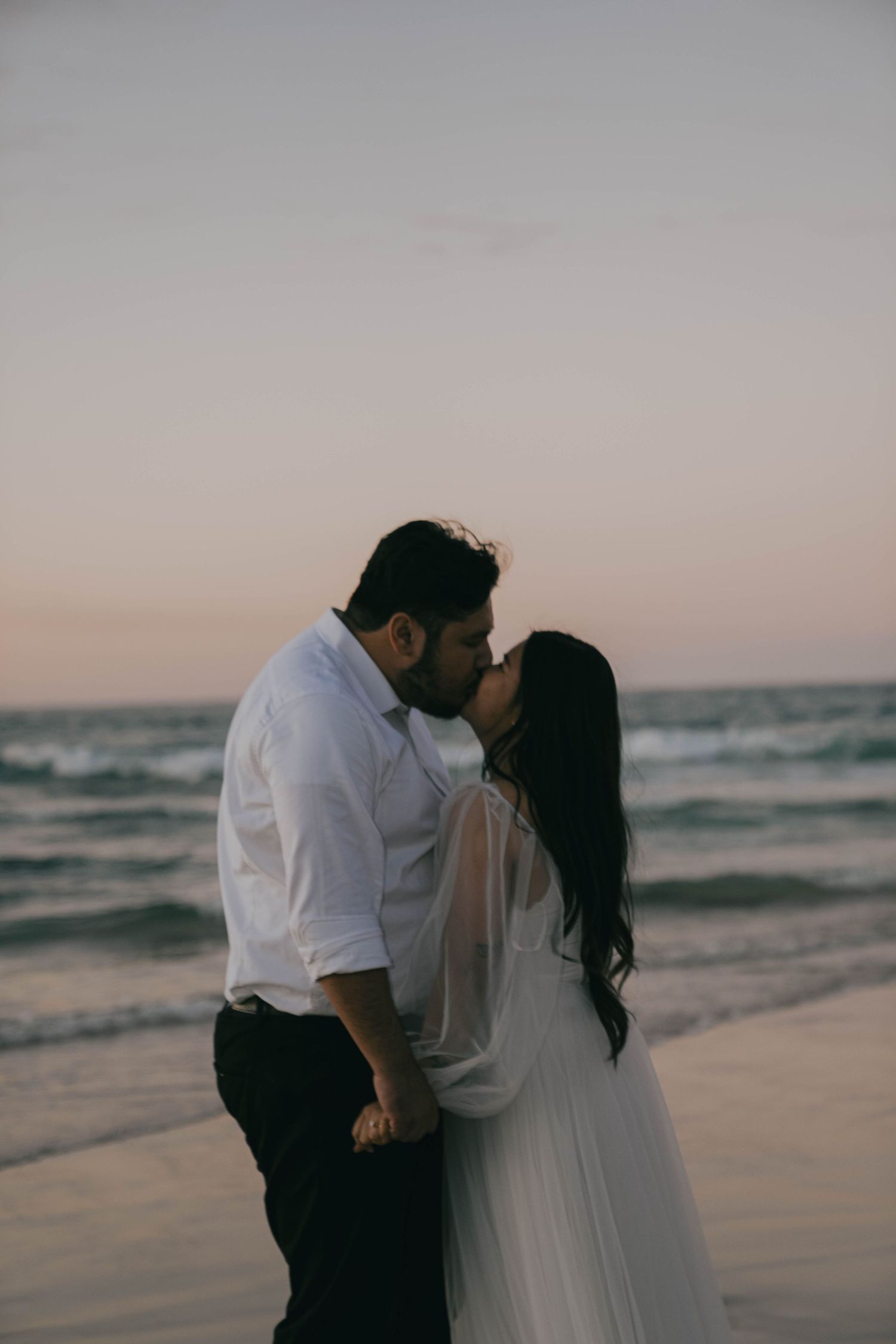 A romantic couple embraces on a serene beach at sunset with ocean waves in the background.