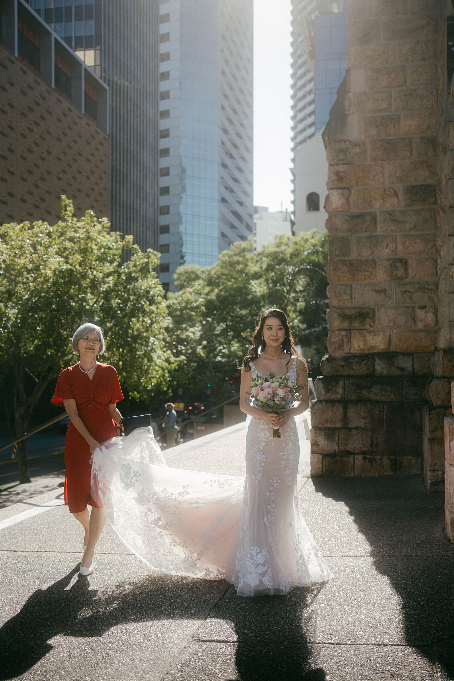A bride in a white lace gown with flowing veil stands on a sunny city street alongside a companion in red.