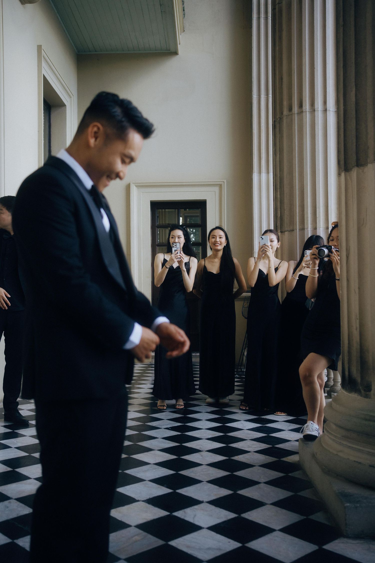 People dance on a dramatic black and white checkerboard floor in formal attire against white pillars and walls.