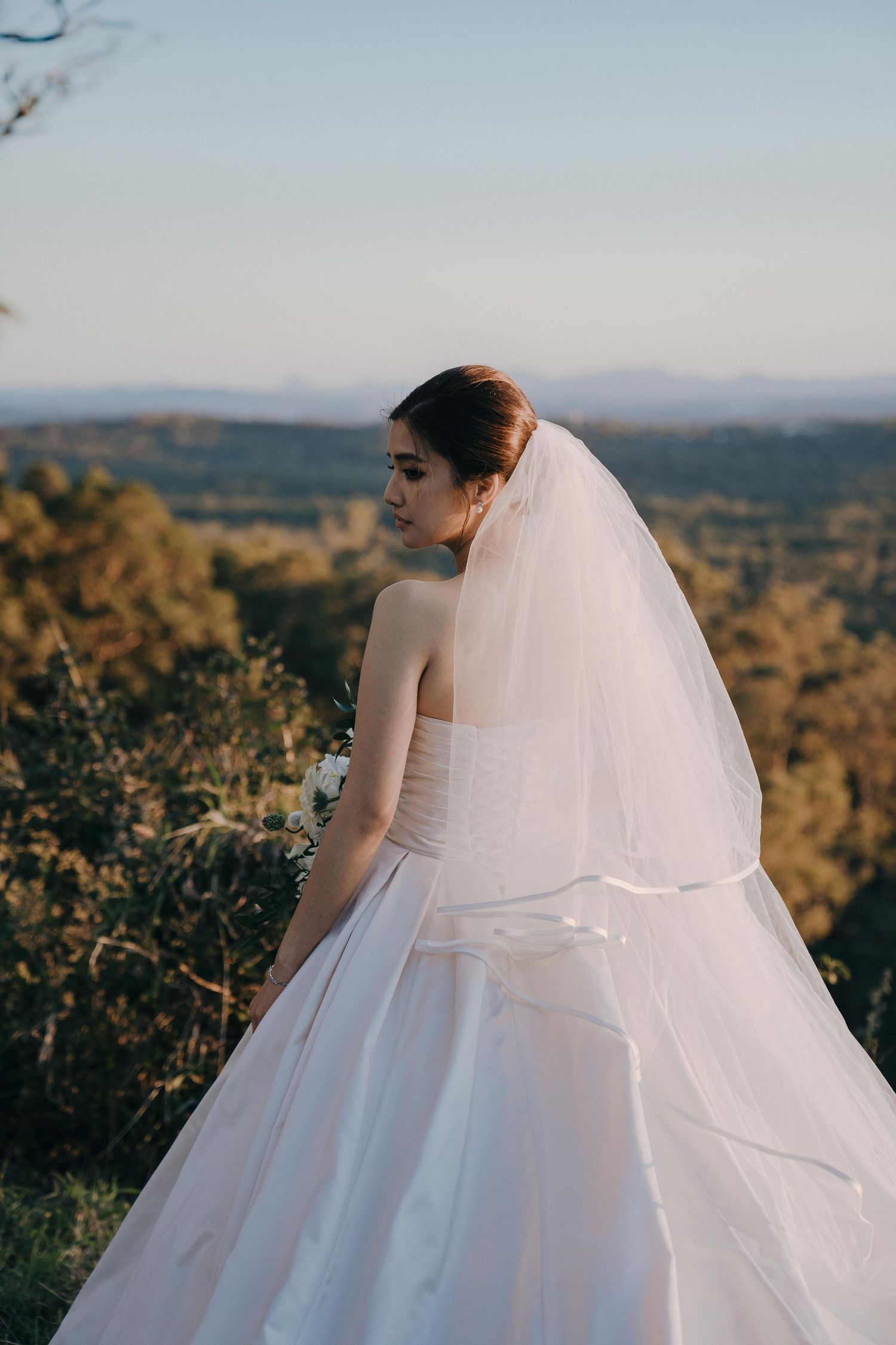 A bride in a flowing white ballgown and veil poses against a scenic mountainous landscape at sunset.