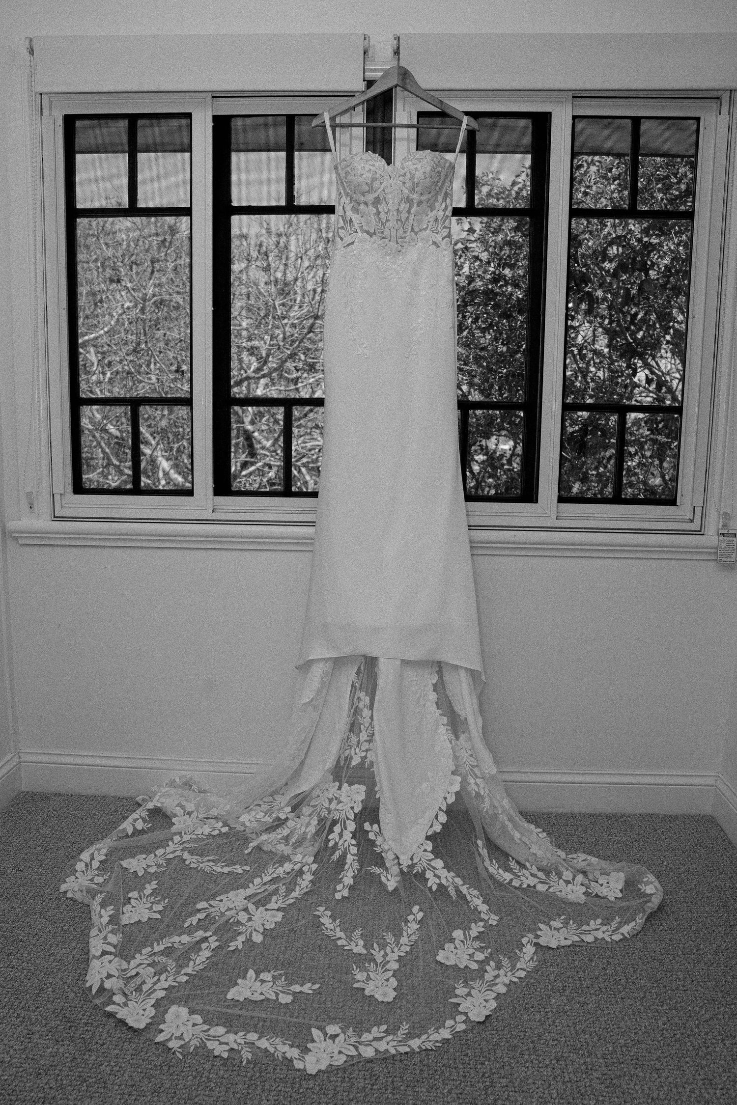 A wedding dress with intricate lace train displayed on a carpeted floor in a room with large windows.