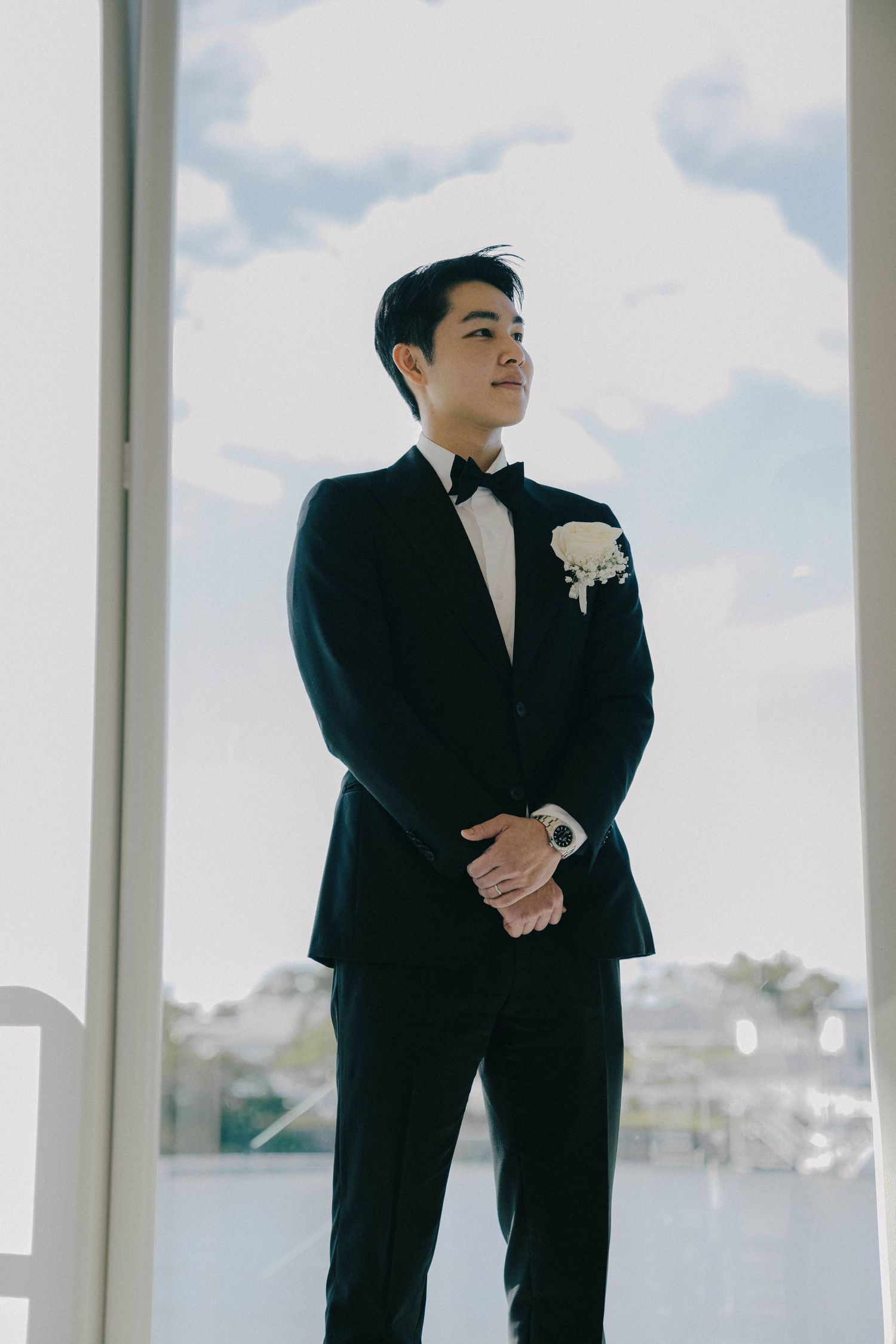 A person in a black suit and white boutonniere stands in a bright modern space with large windows.